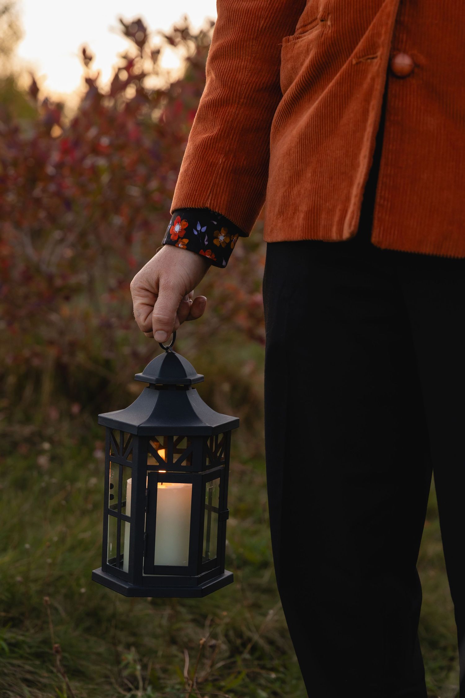 A person in an orange coat holds a black decorative lantern with candlelight glowing inside at dusk.