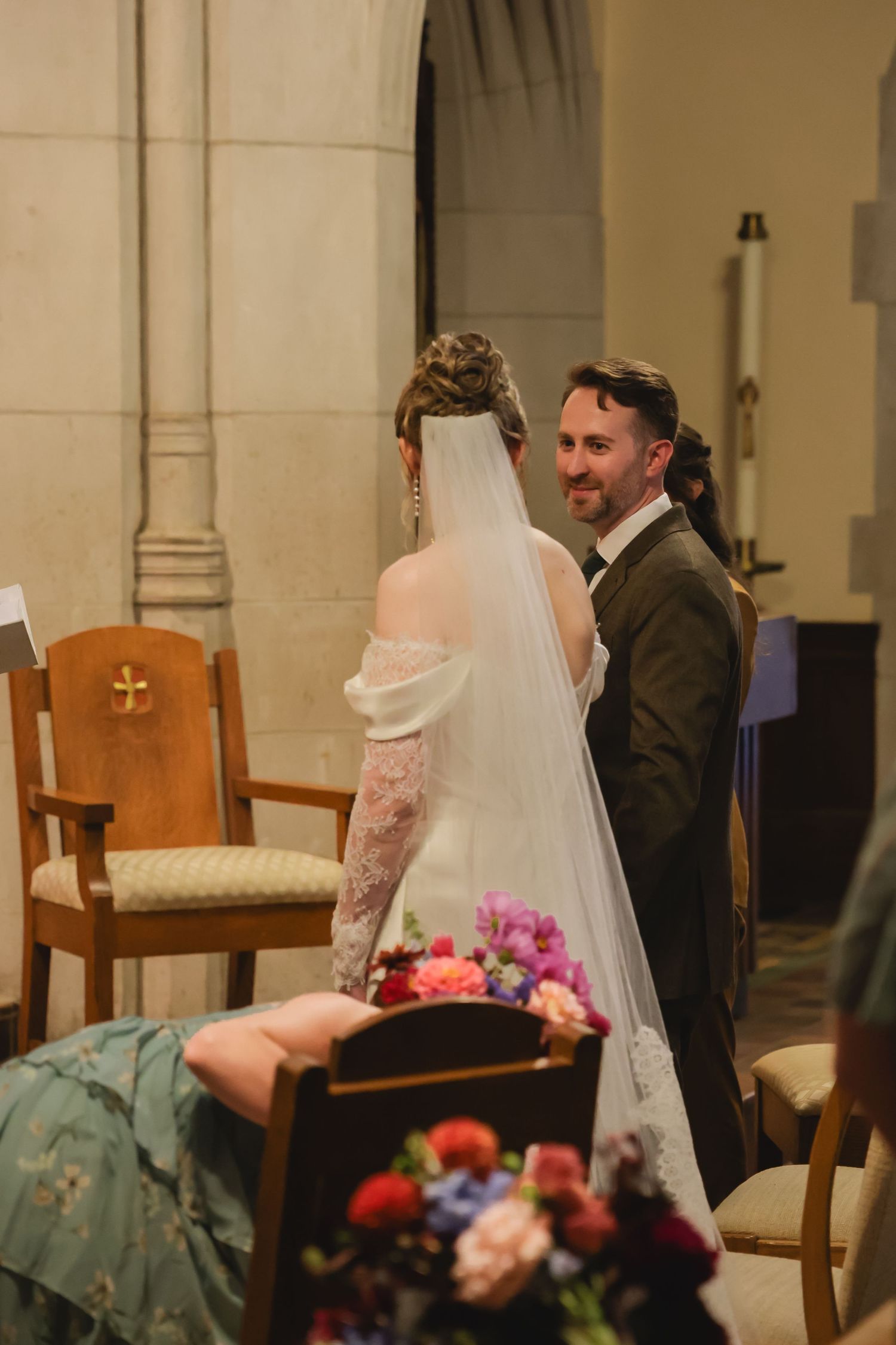 A couple sits in wooden chairs during a wedding ceremony in a church with soft lighting and floral decorations.
