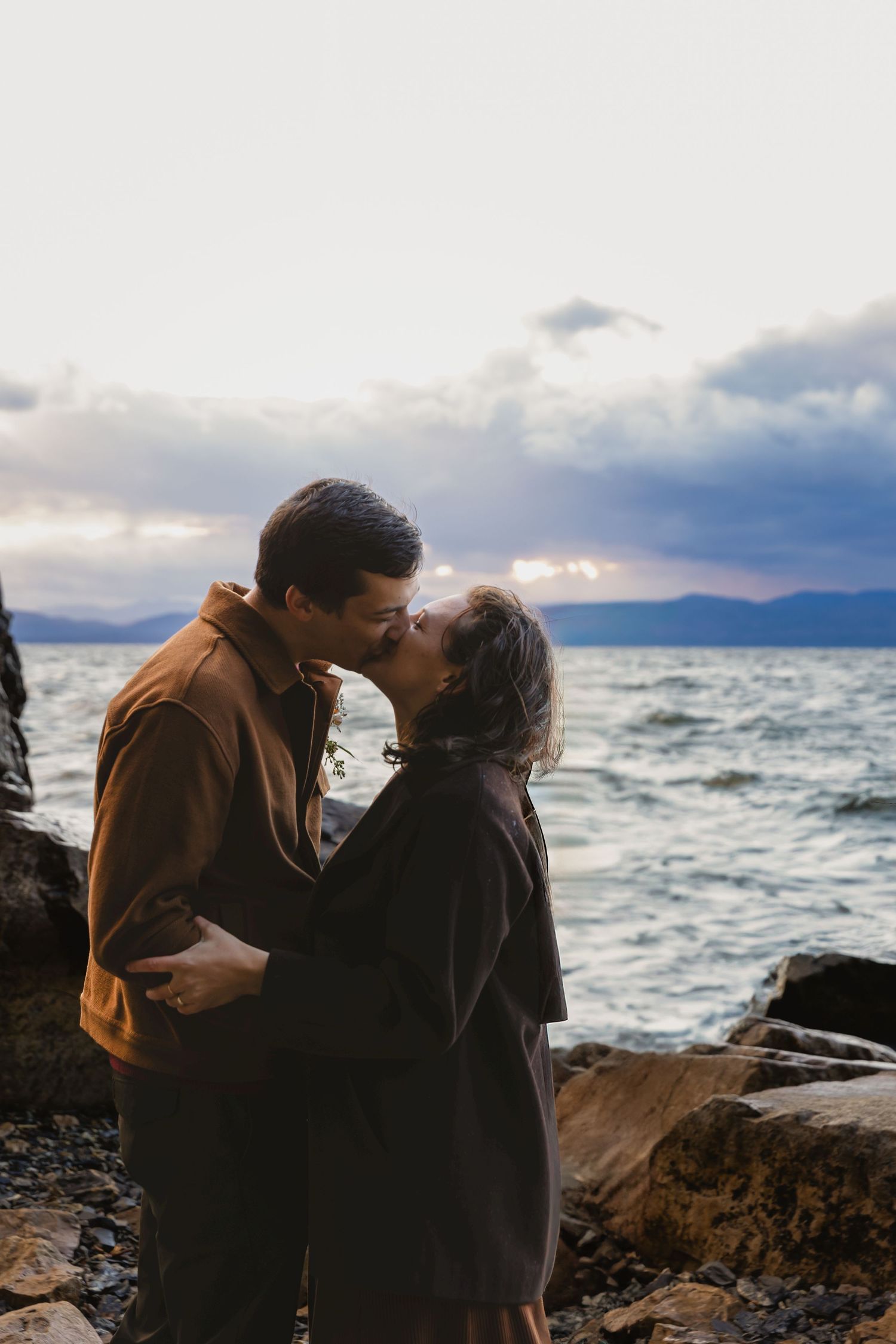 Romantic couple embraces on rocky shore with stormy waters and mountains in background during moody sunset.