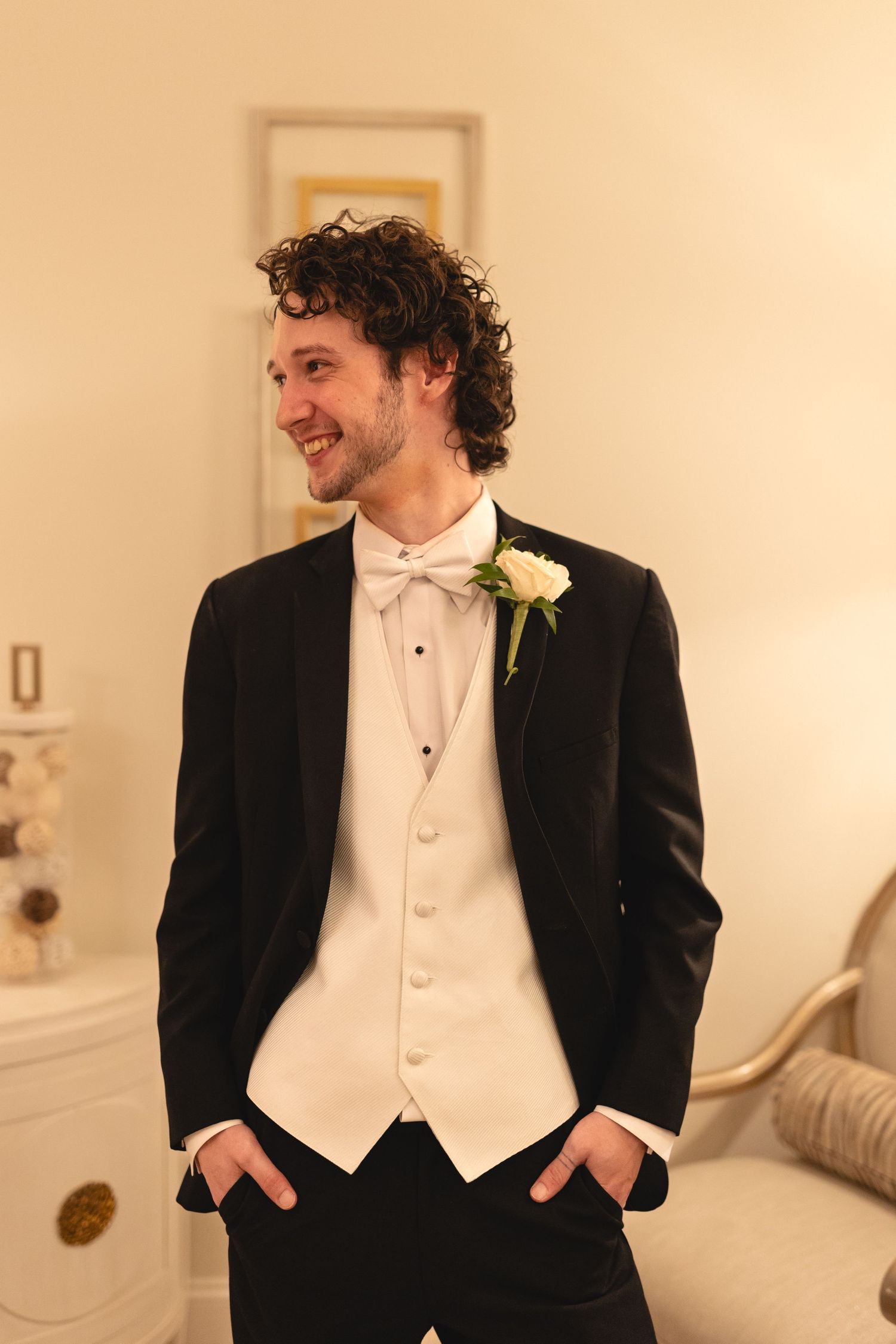 Groom wearing black tuxedo with white vest and bowtie smiles while standing with hands in pockets at wedding.