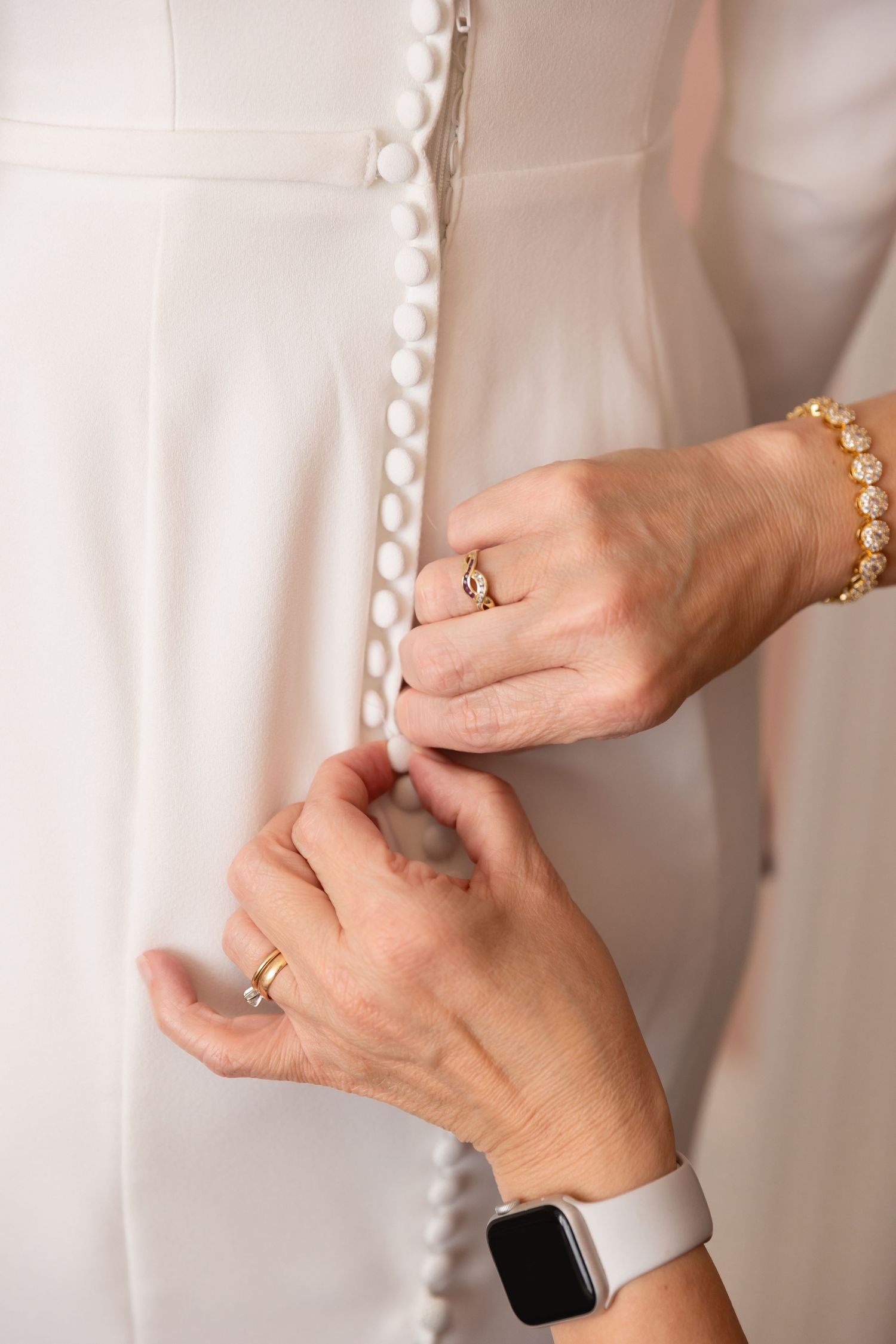 Hands button up pearl closures on a white dress while wearing gold jewelry and a modern smartwatch.
