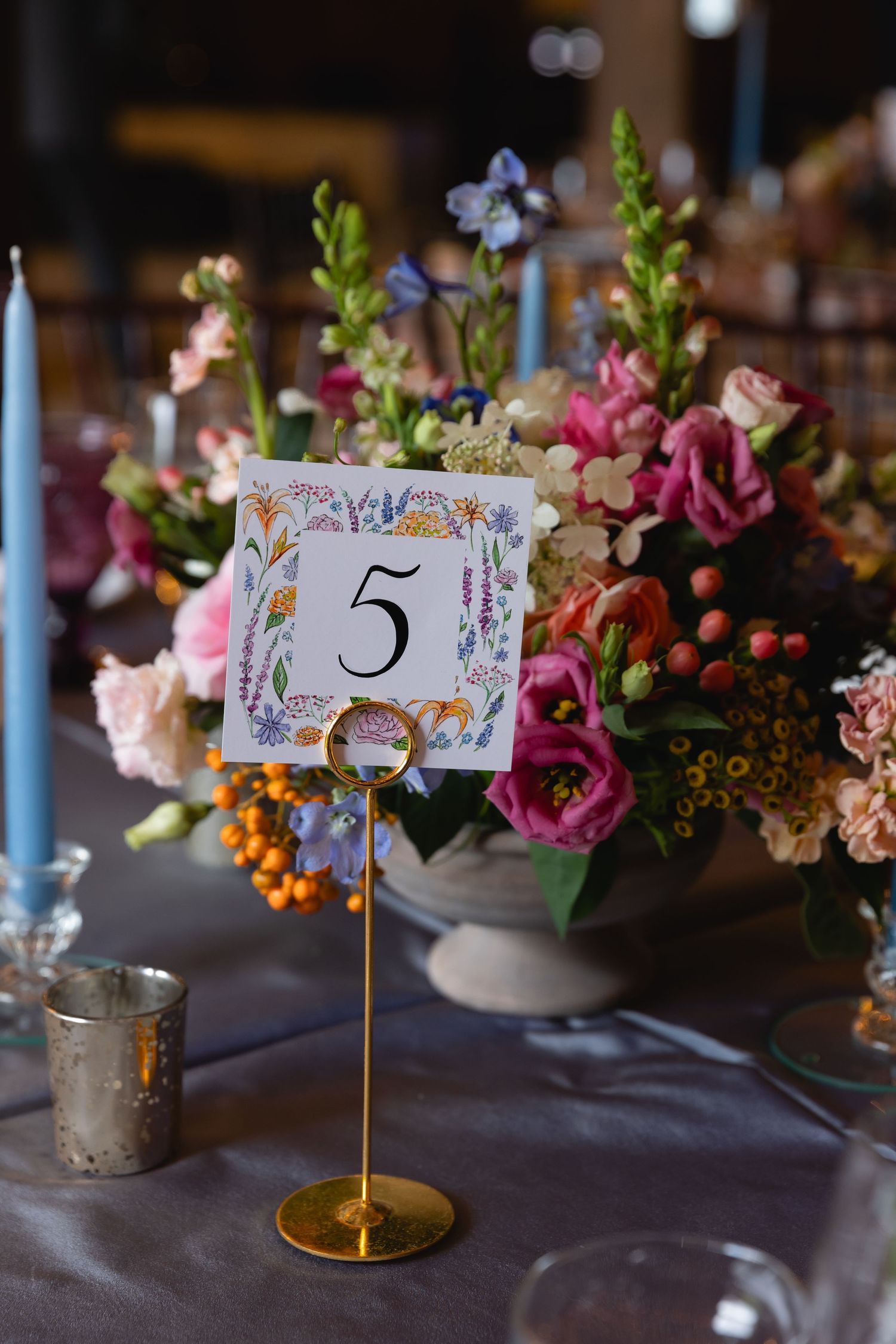 An elegant table setting with floral centerpiece, blue candles, and decorative table number 5 on a gold stand.