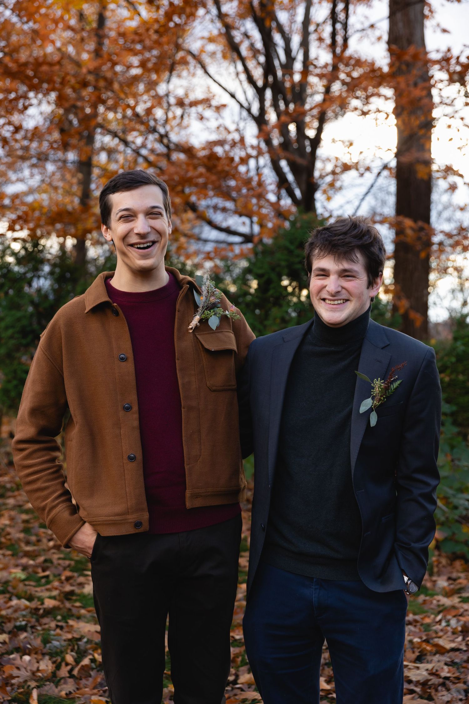 Two people in fall outdoor setting wearing sweaters and boutonnieres with autumn foliage in background.