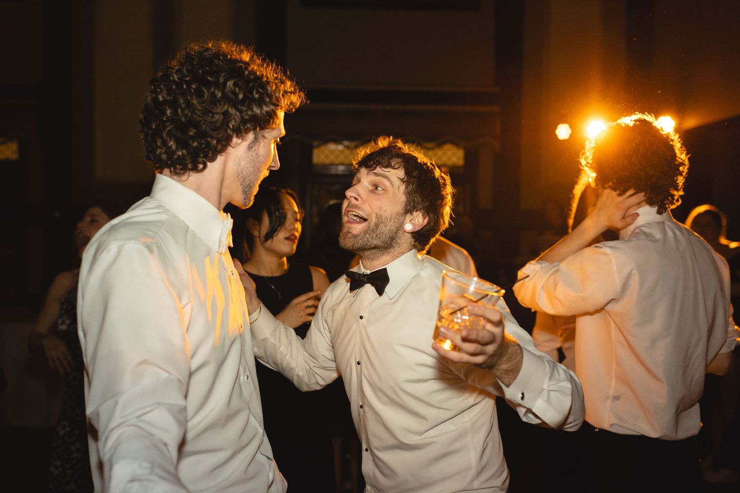 Two people in formal white attire dancing and laughing together at an evening celebration with warm lighting.
