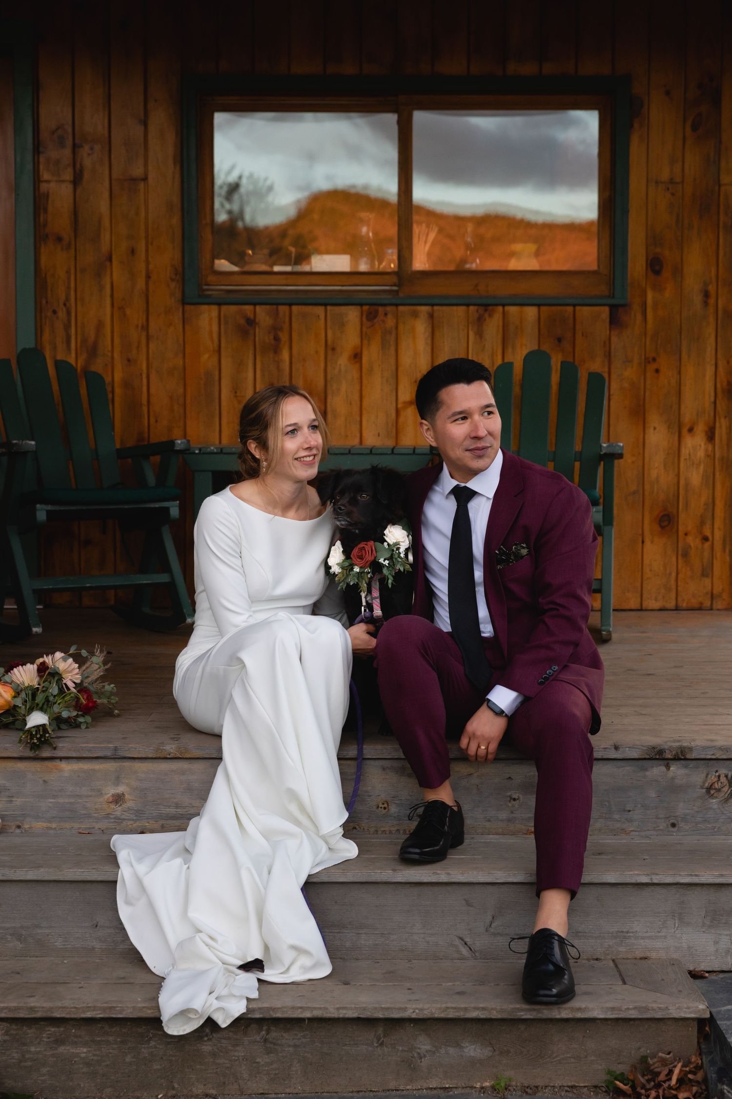 A couple sits on a wooden porch wearing wedding attire - a white dress and burgundy suit - in front of mountain scenery.