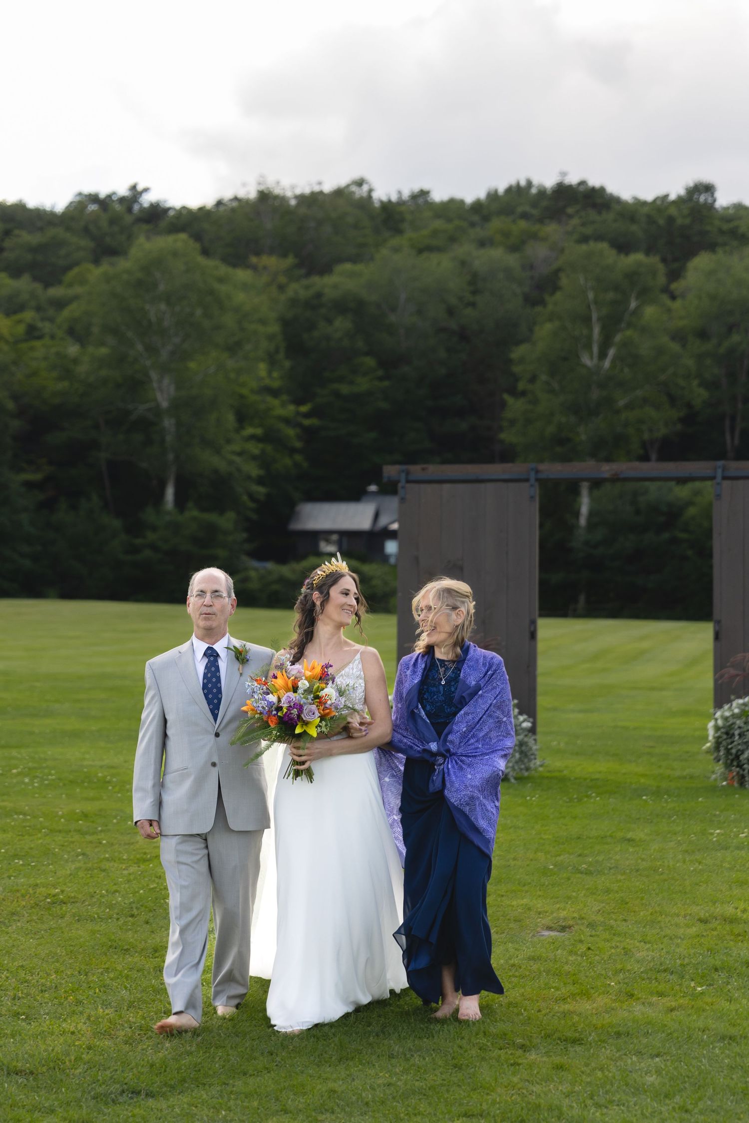 Three wedding guests stand together on a lush green lawn with trees in the background.