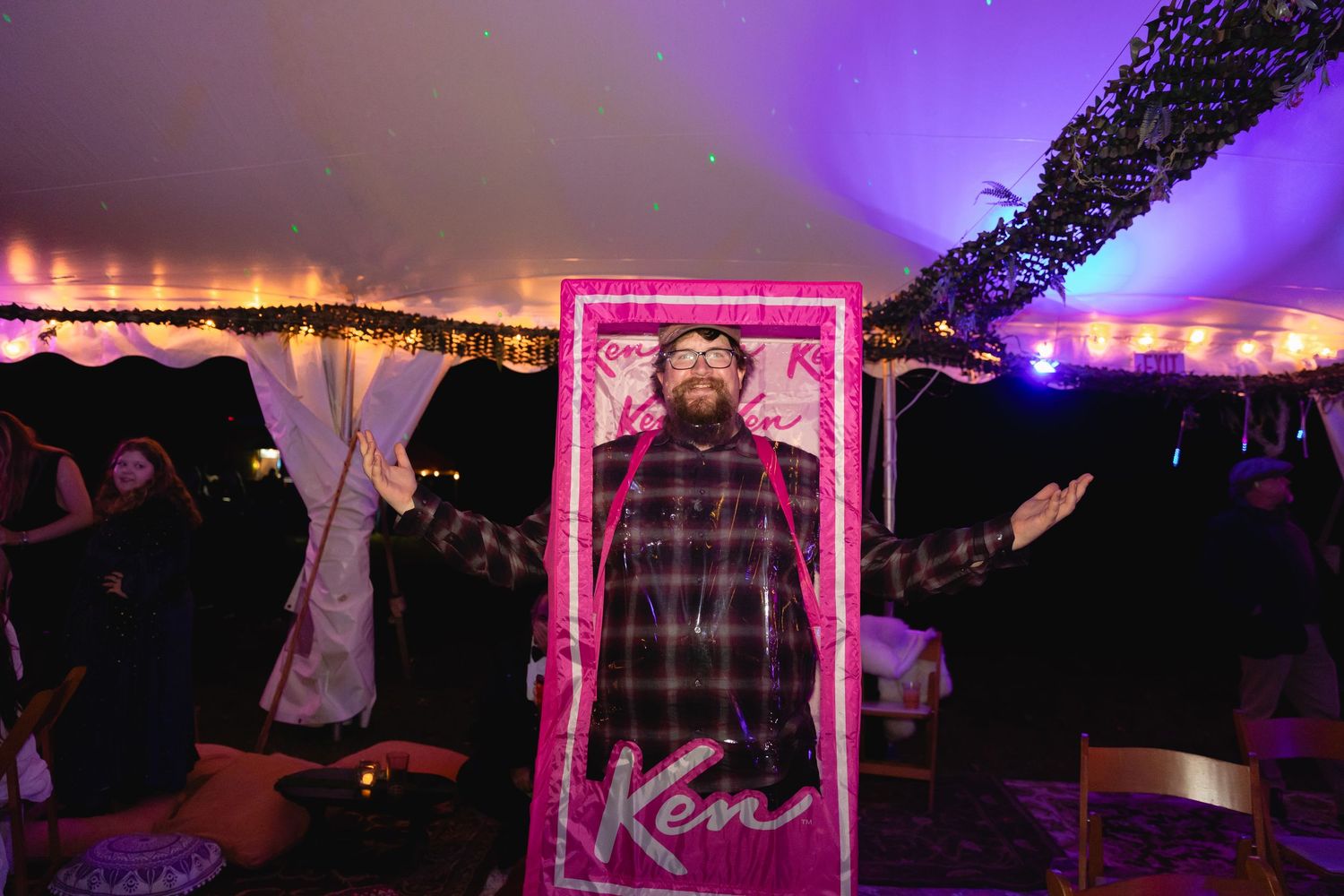 Bearded man posing inside a neon pink Ken-themed photo frame at a wedding reception.