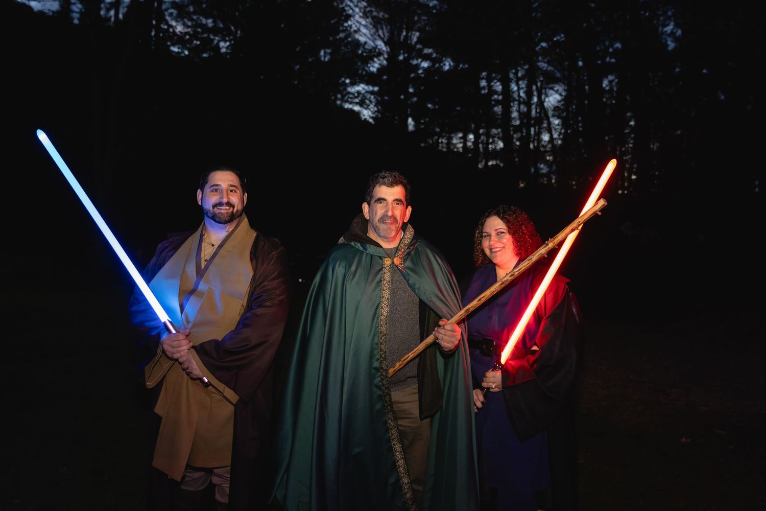 Three wedding guests in fantasy costumes posing with glowing lightsabers in a dark forest setting.