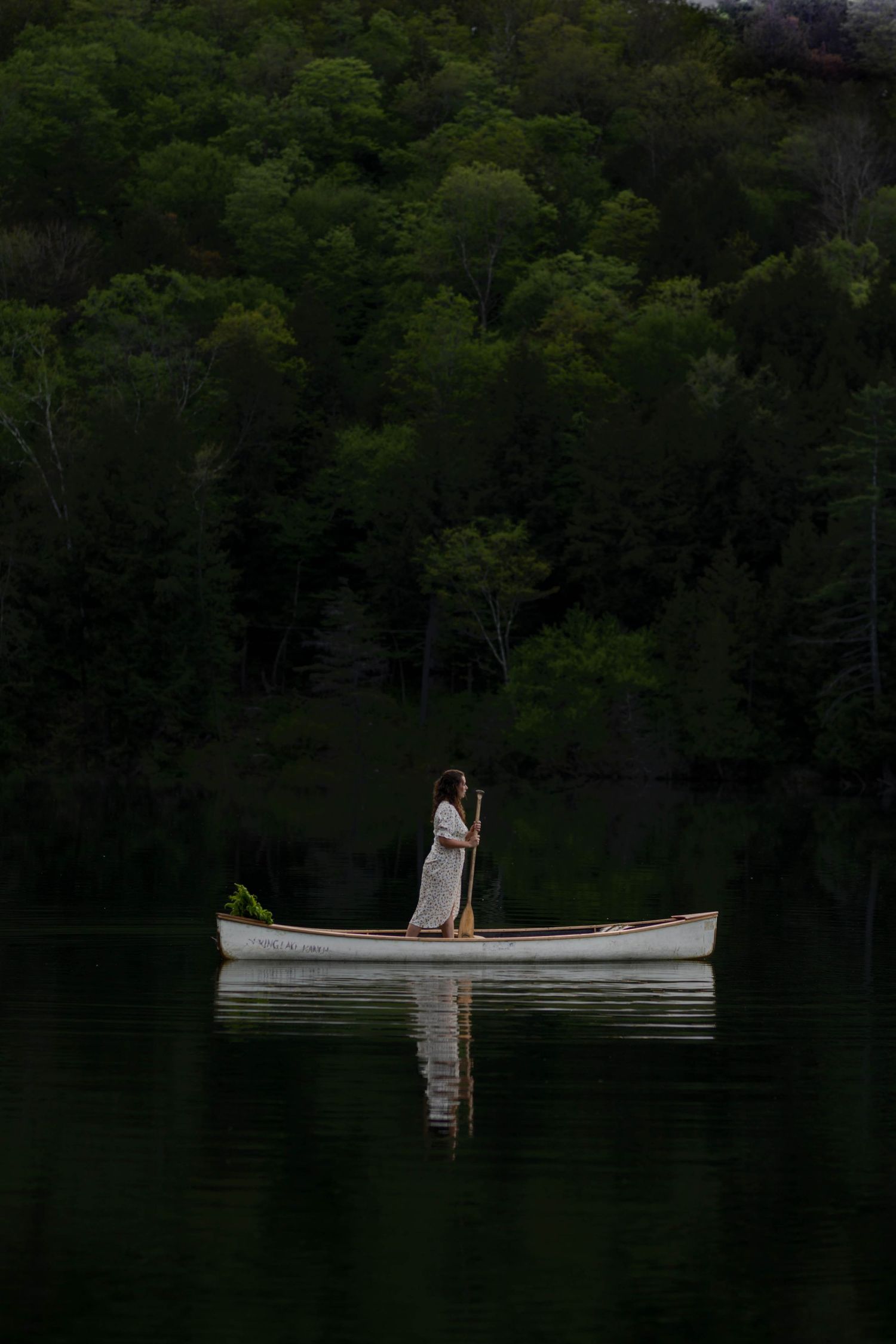 A woman in a white dress stands paddling a canoe on a calm lake with lush green forest behind her.