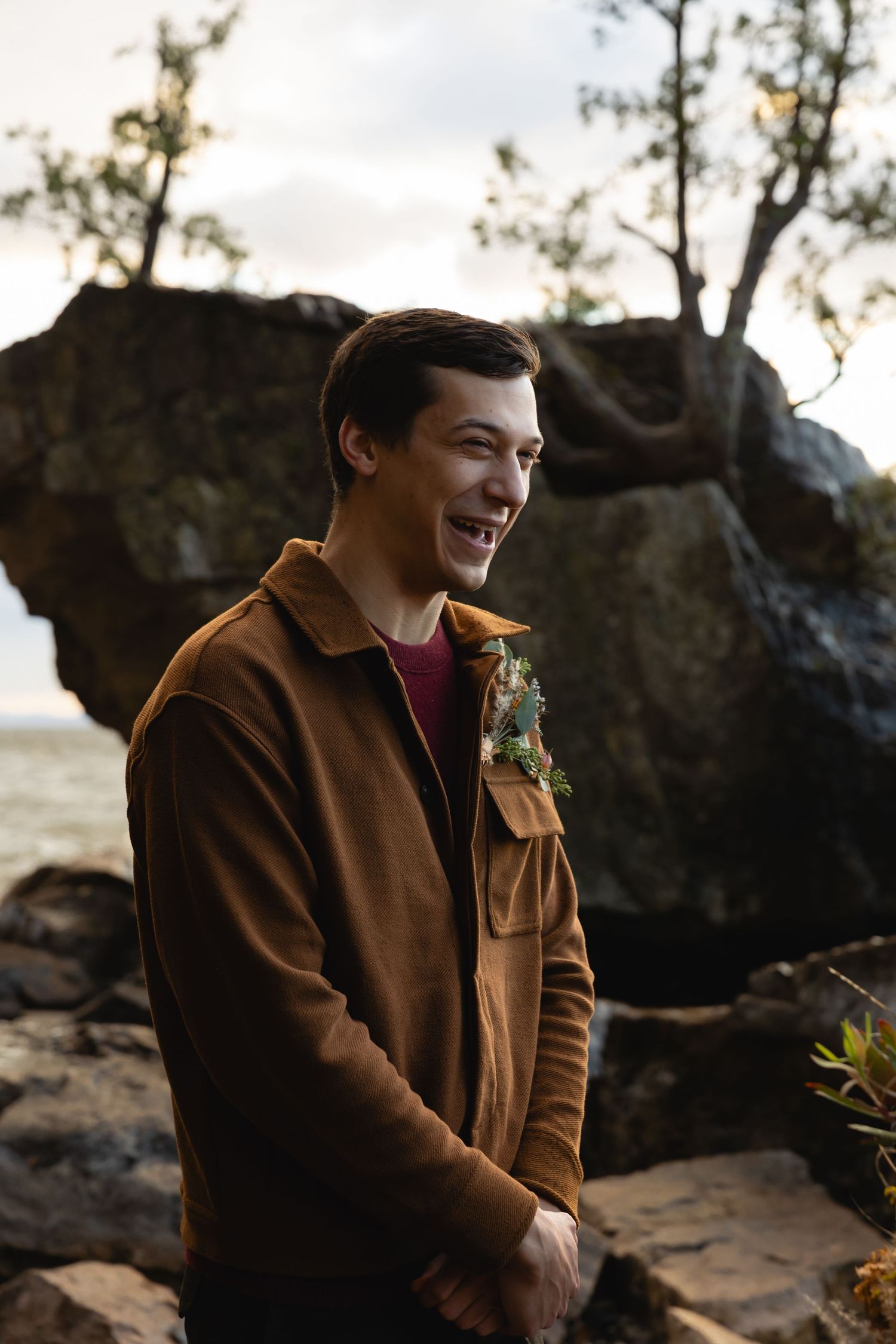 A person in a brown jacket laughs joyfully while standing near large rocks in nature during golden hour lighting.