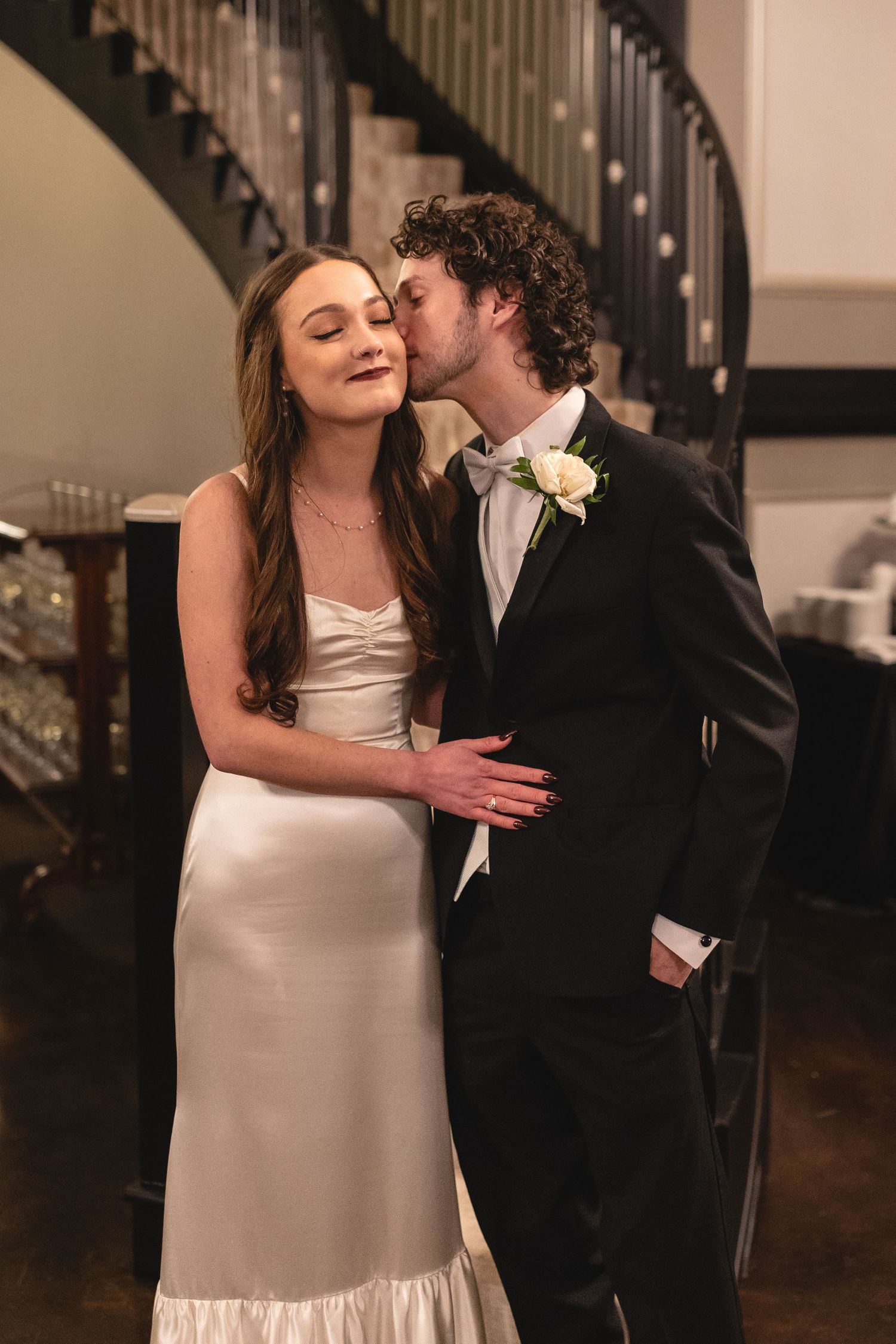 A newlywed couple shares a romantic moment in front of an elegant staircase, the bride in white and groom in black tuxedo.