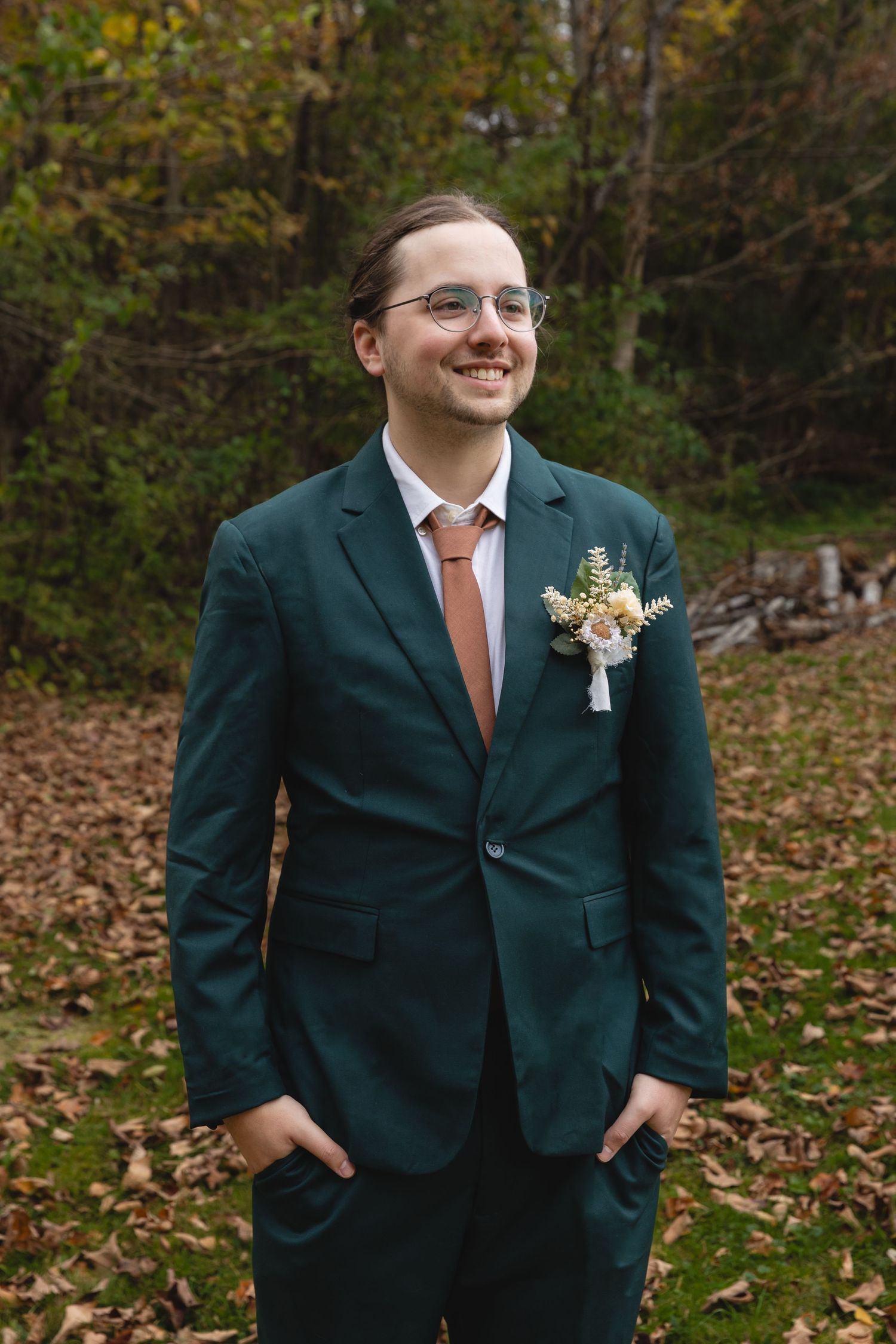 Person in green suit jacket with boutonniere smiling in autumn forest setting.