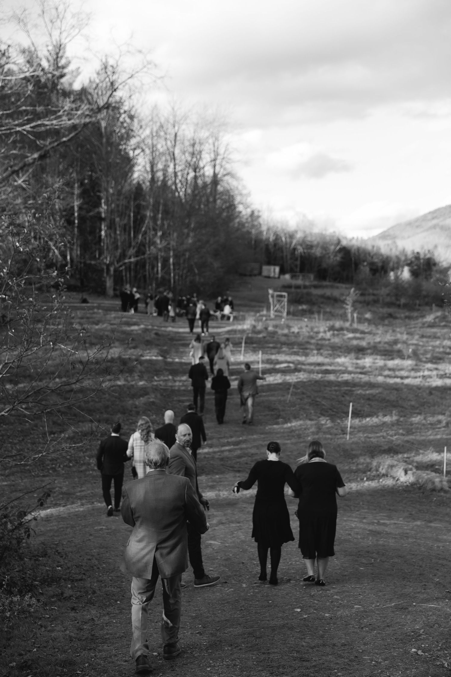 A black and white photograph shows people walking along a rural hillside path through bare trees on an overcast day.