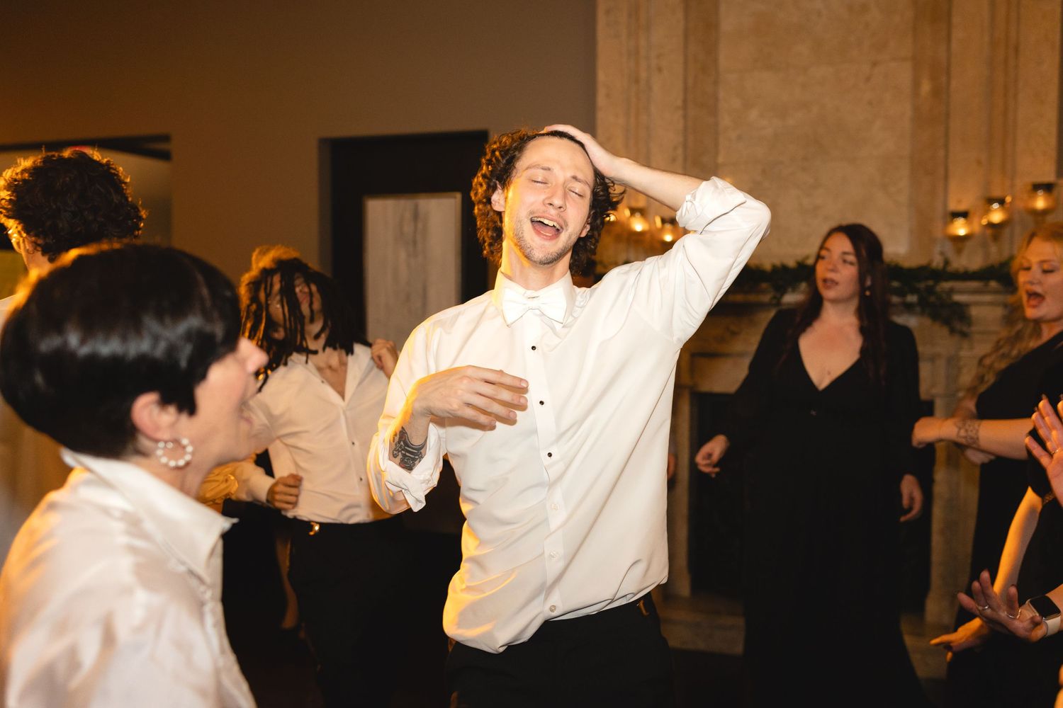 A lively dance scene at a wedding reception shows guests in white shirts dancing and laughing under warm lighting.