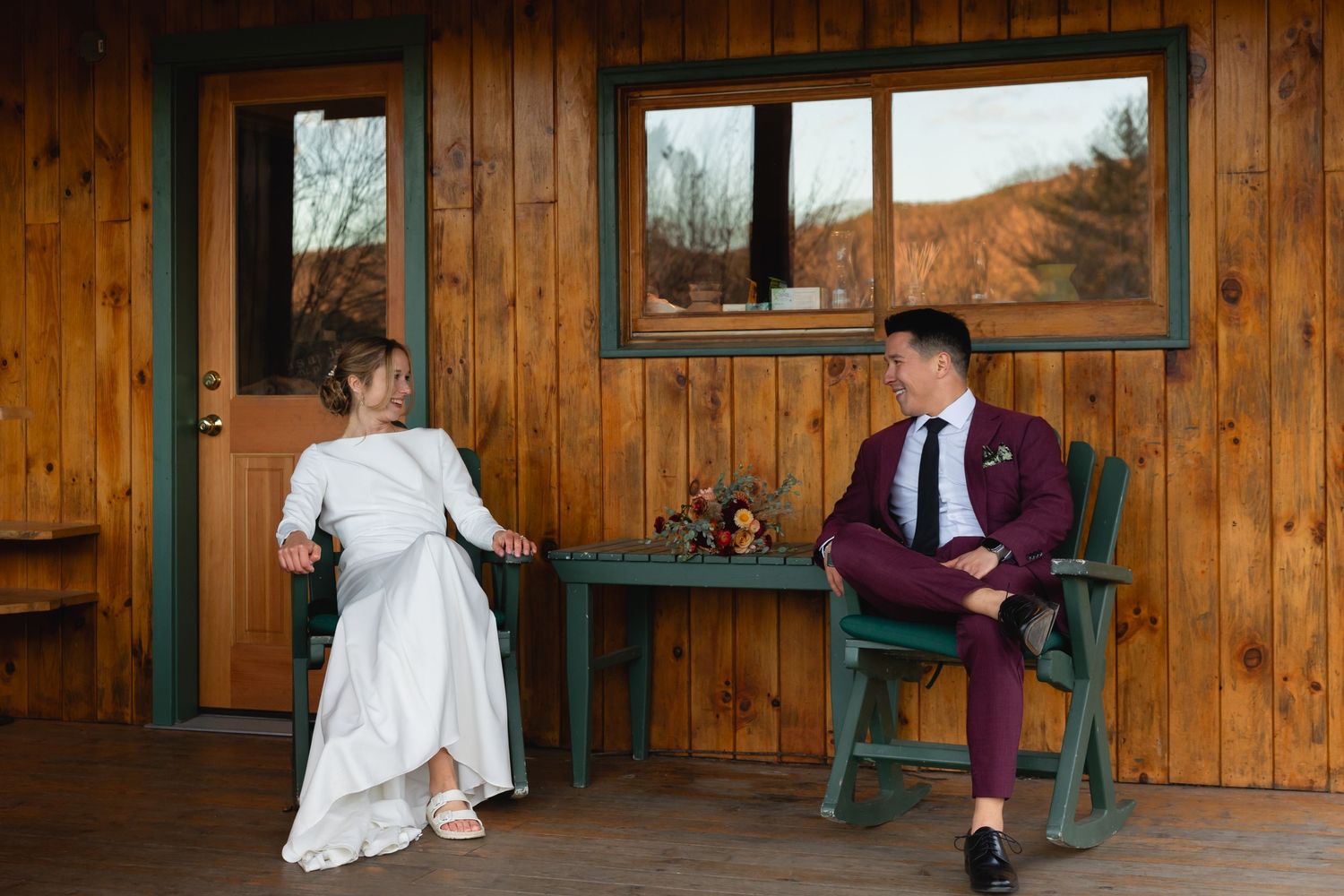 Couple sits together on rustic wood cabin porch, wearing wedding attire on an autumn day.