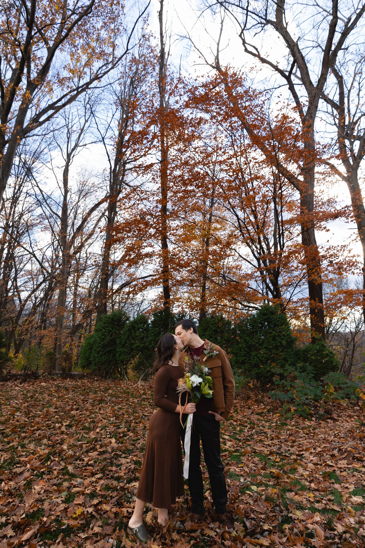 A couple in fall attire embrace among autumn trees with fallen leaves scattered on the ground in a woodland setting.
