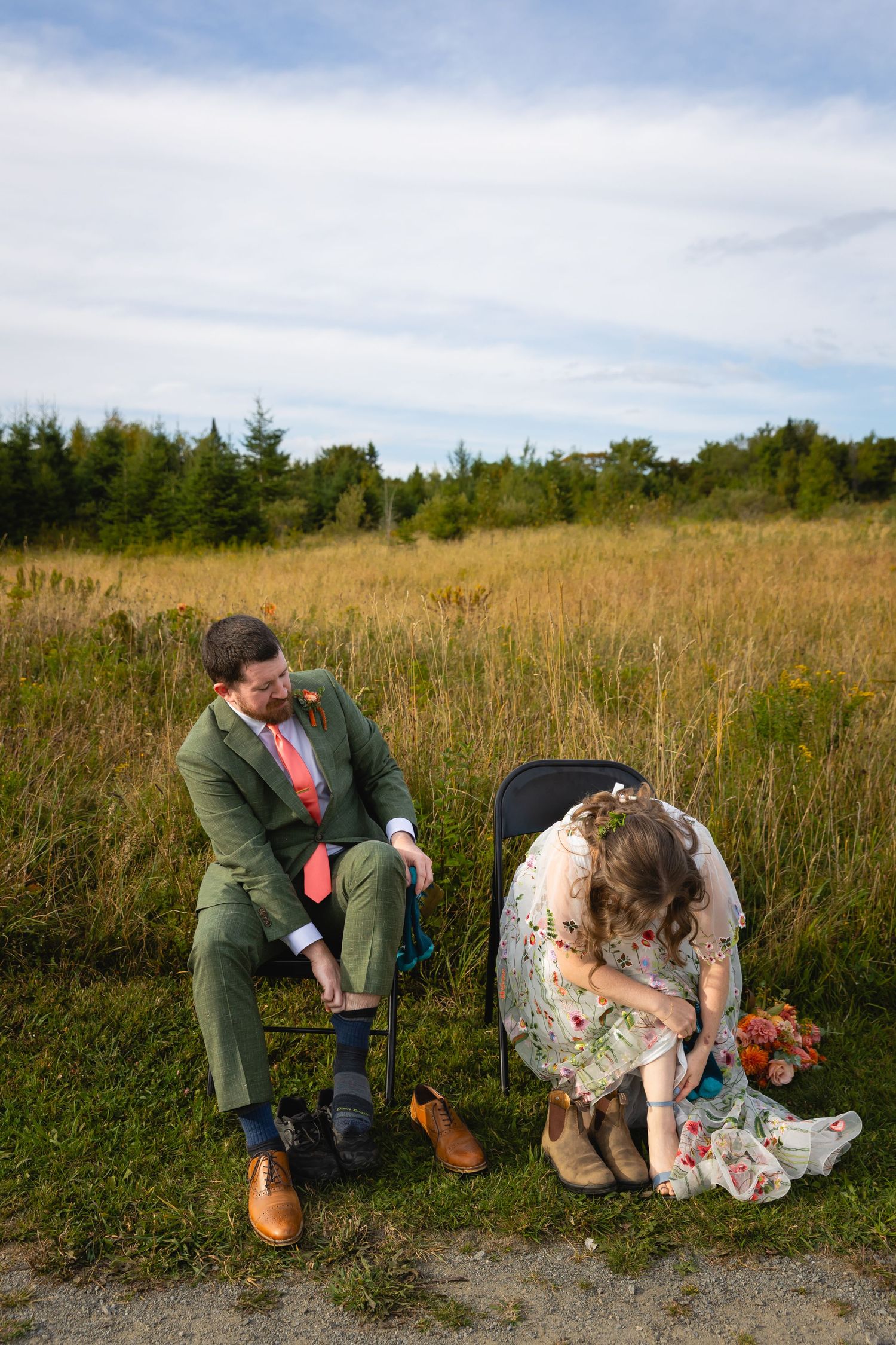 A person in green attire crouches down to inspect a large mushroom growing in a grassy field on a cloudy day.