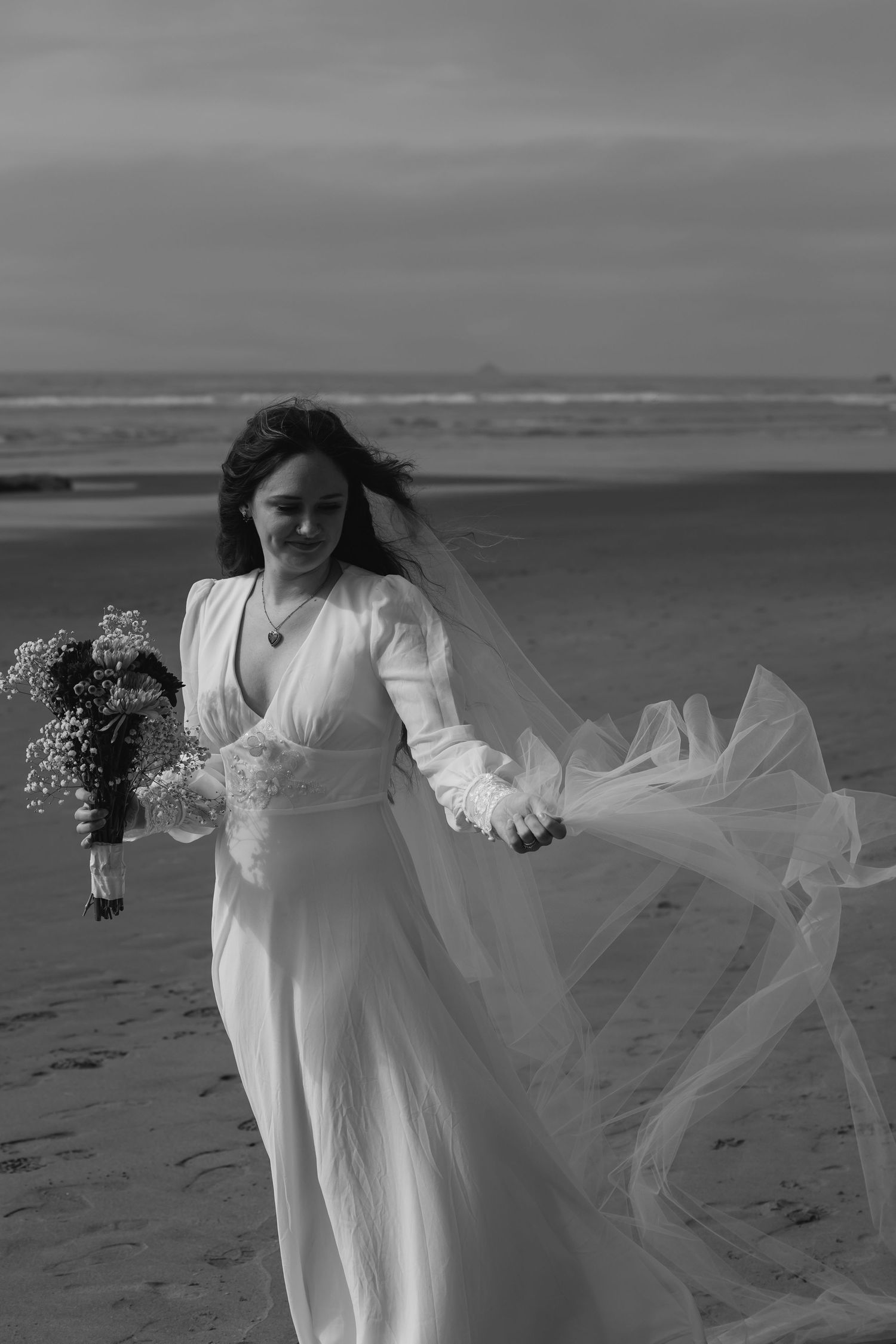 A figure in a flowing white dress stands on a beach at sunset holding flowers as fabric billows in the ocean breeze.
