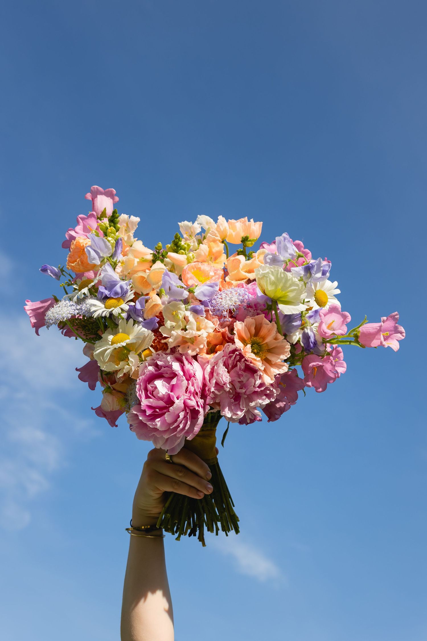 A hand holds up a vibrant bouquet of pink, peach and white flowers against a bright blue sky background.