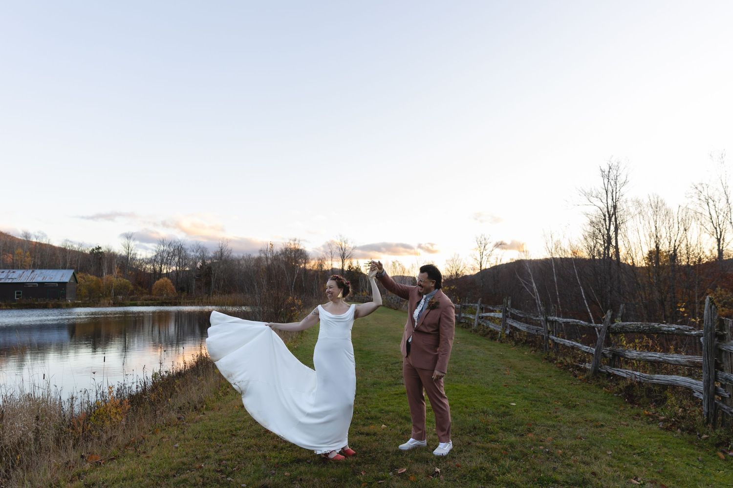Couple poses playfully on grassy path beside lake during sunset wedding photoshoot.