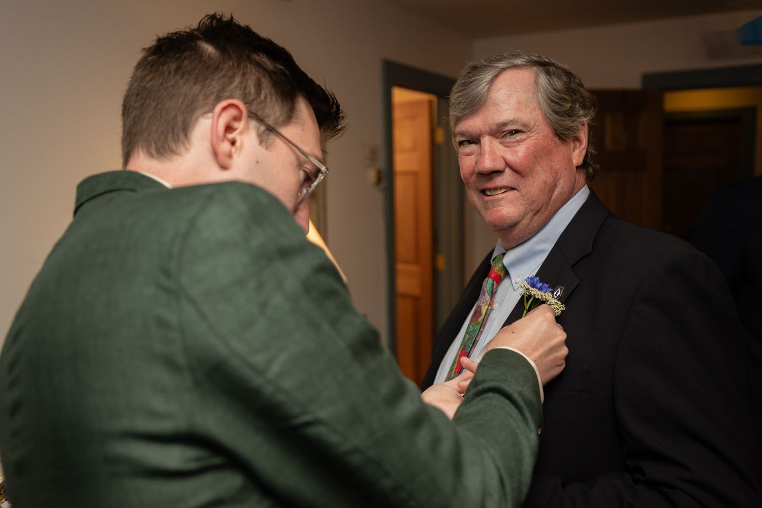 Two people adjust a boutonniere during wedding preparations indoors.
