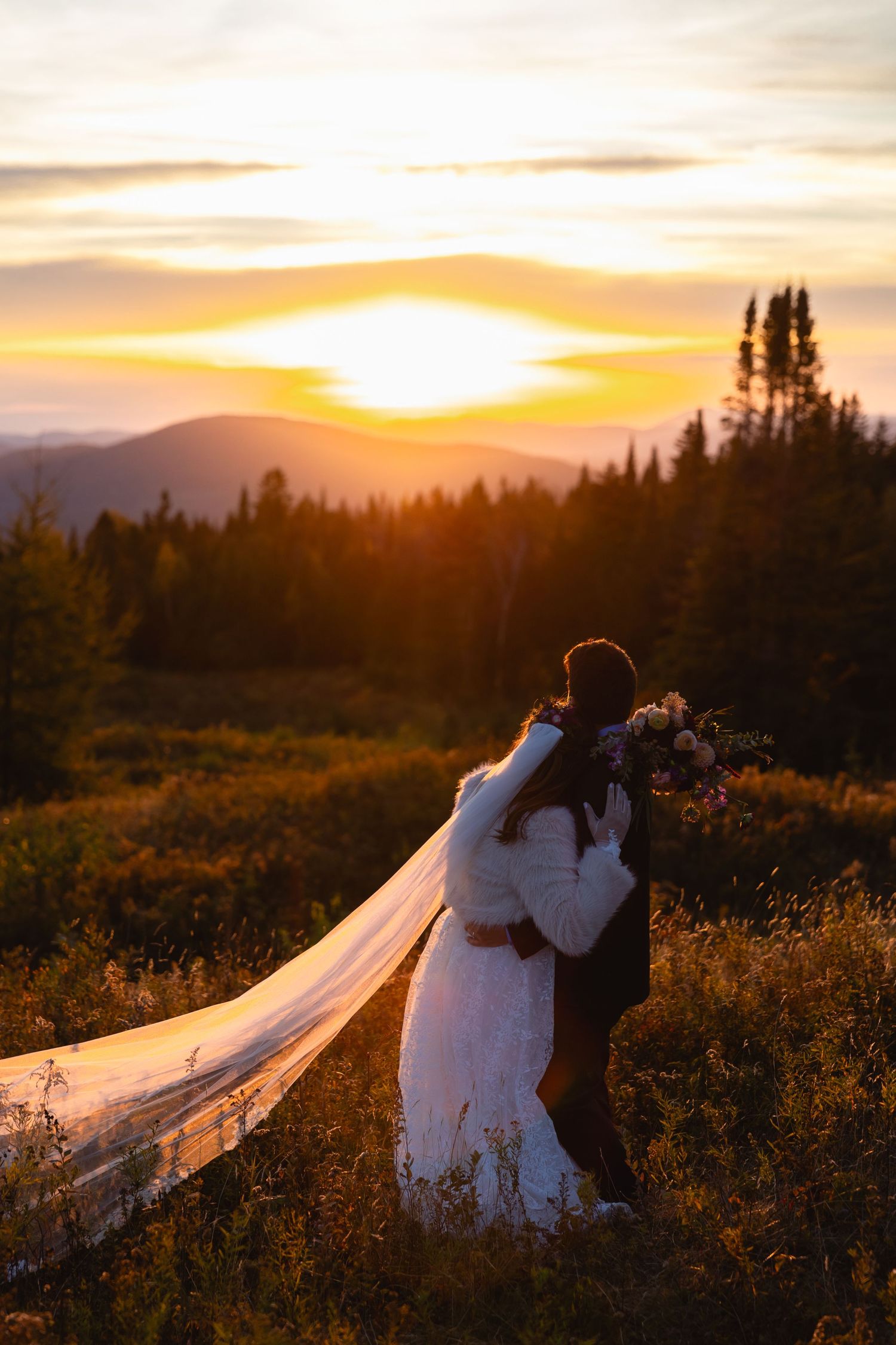 Bride and groom embrace at golden sunset overlooking mountain landscape, bridal veil flowing in wind.