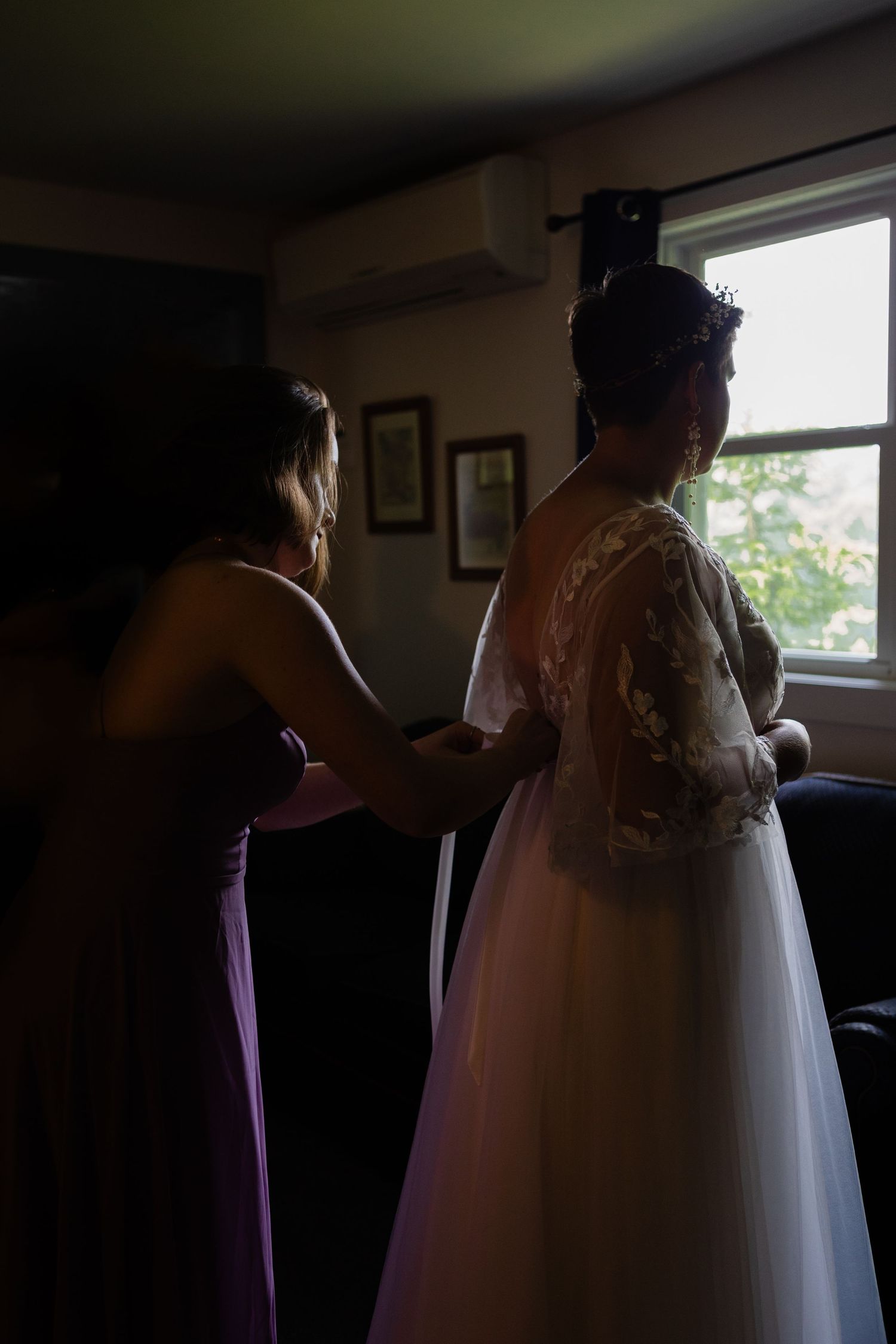 Helping with wedding dress preparations in shadowy room by window.