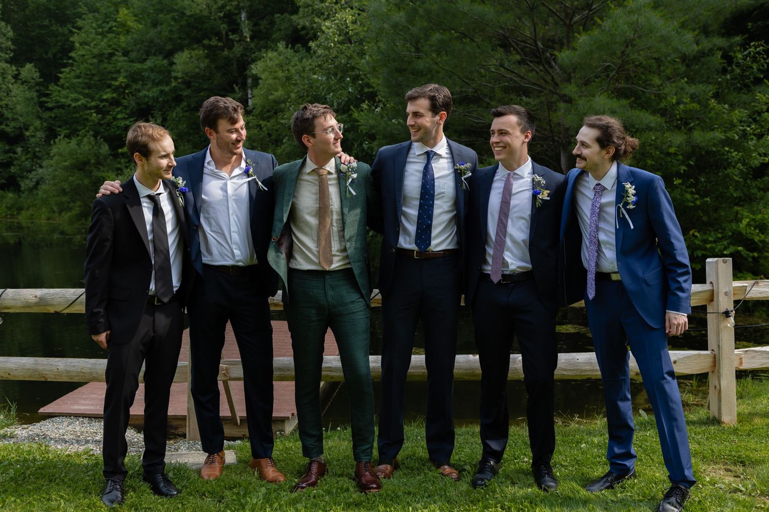 A group of groomsmen in suits pose together outdoors in front of a wooden fence.