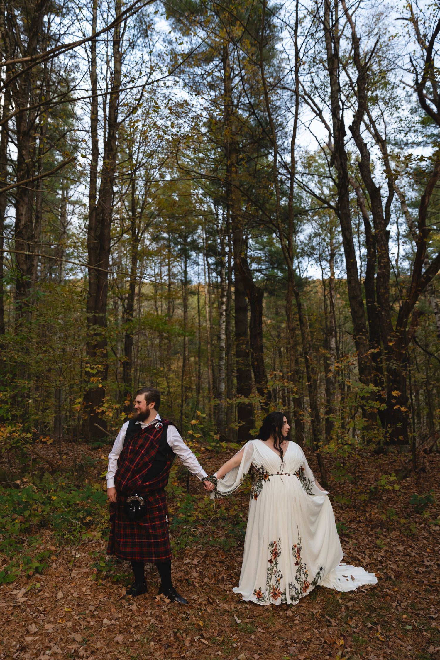 A couple in Celtic wedding attire holds hands while walking through an autumn forest.