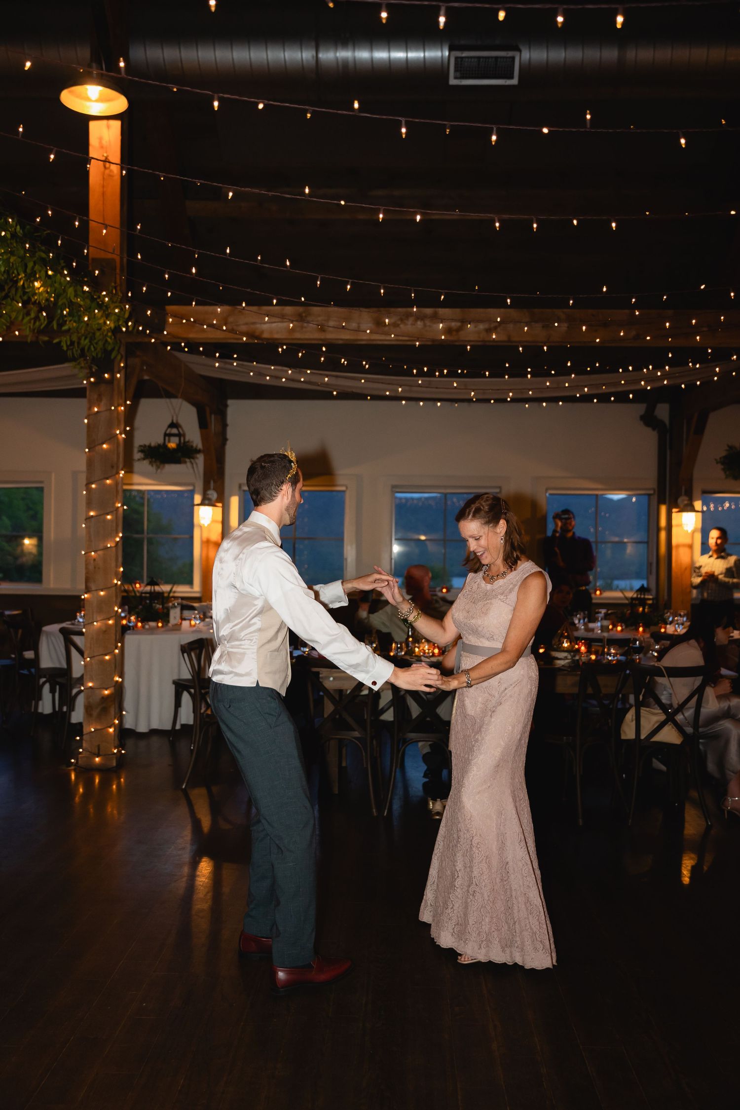 A couple dancing in an elegant reception hall decorated with string lights and wooden beams.