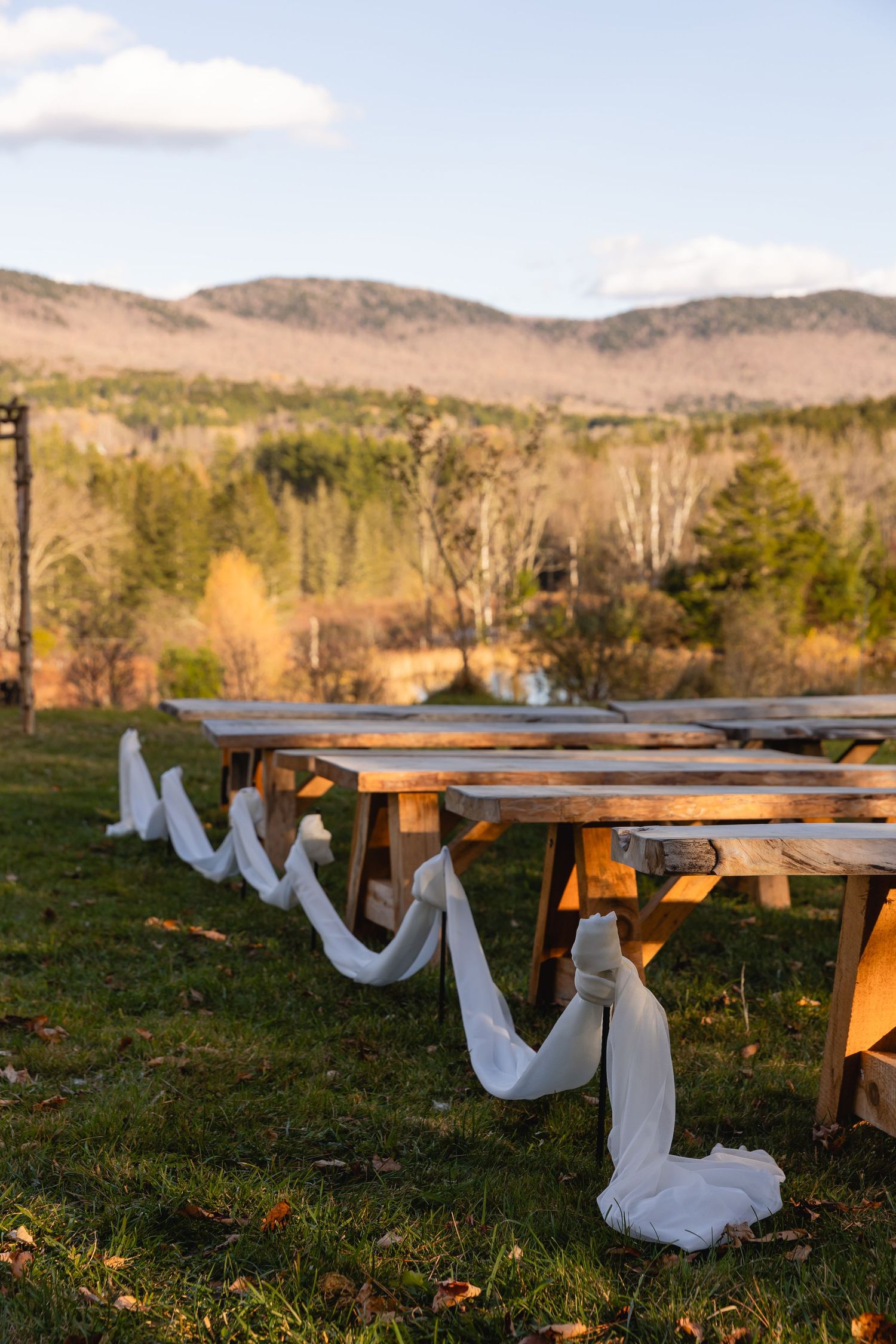 Wooden picnic tables decorated with flowing white fabric drape against a mountainous landscape at sunset.