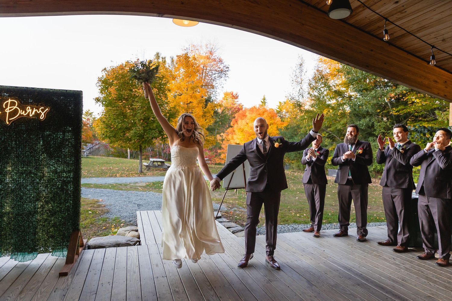 A couple celebrates with wedding party members on a deck with autumn foliage in the background.