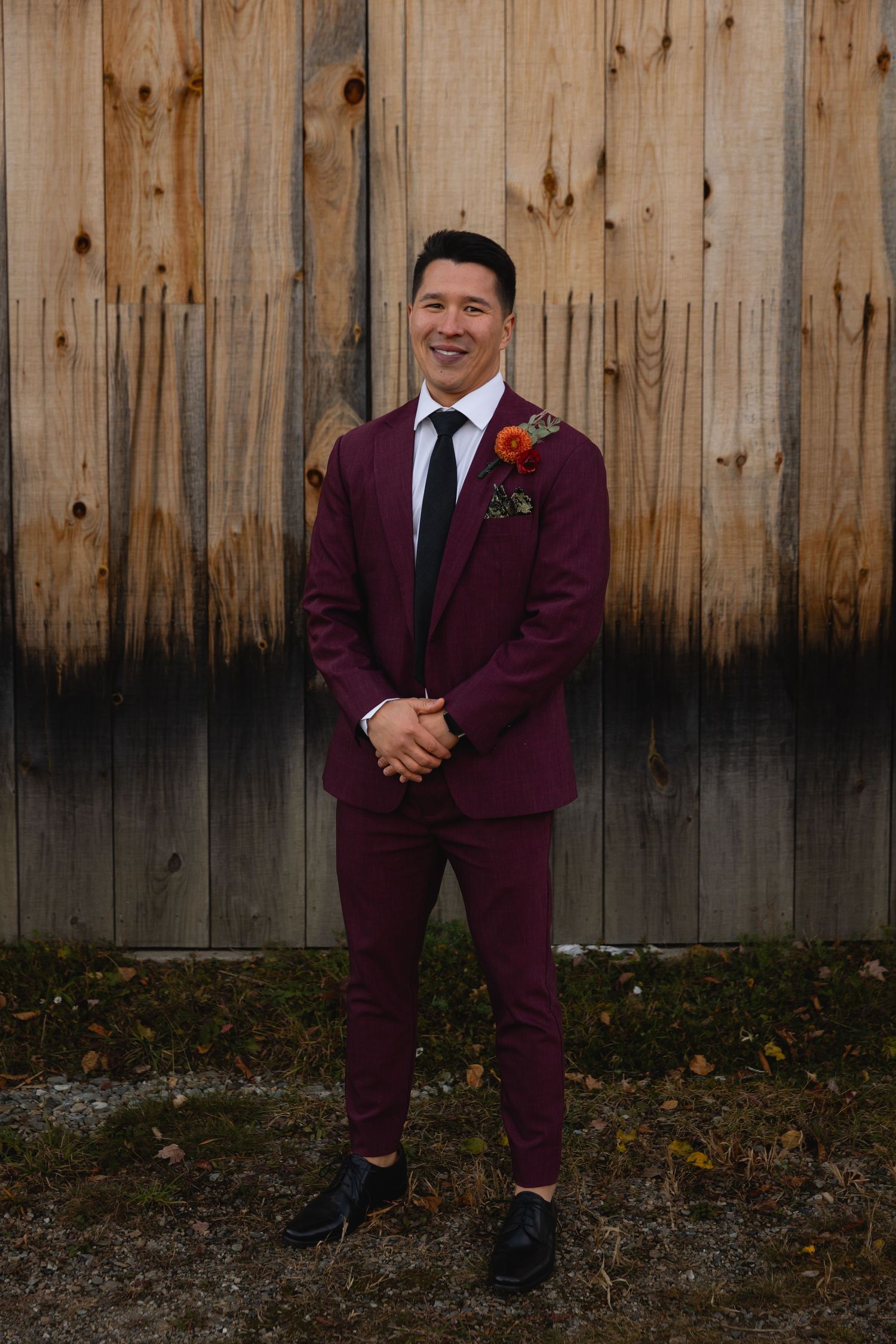 Someone stands confidently in a burgundy suit with a boutonniere against a rustic wooden fence background.