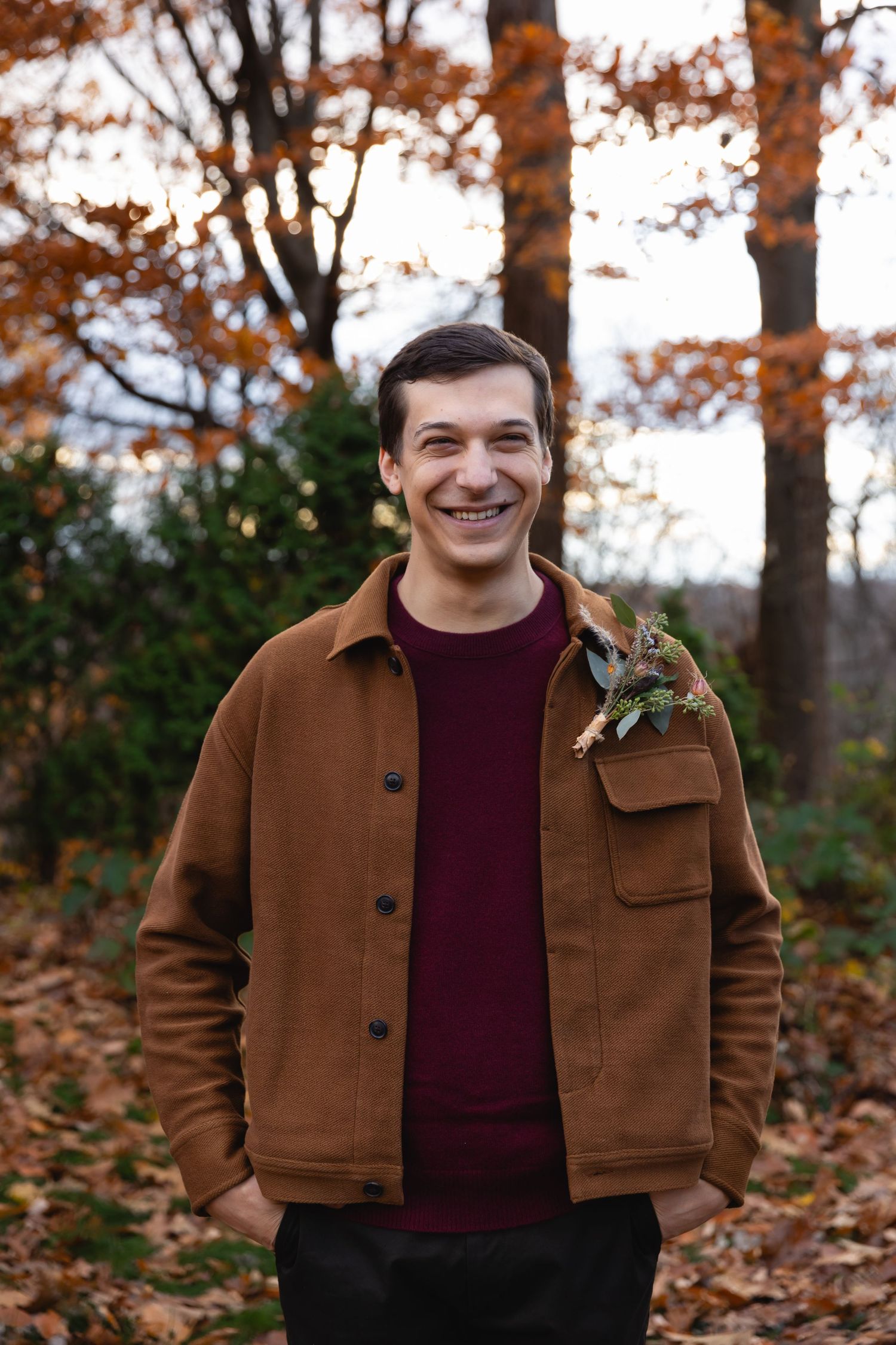 Someone in a brown jacket and burgundy sweater stands smiling among autumn trees with fallen leaves on the ground.