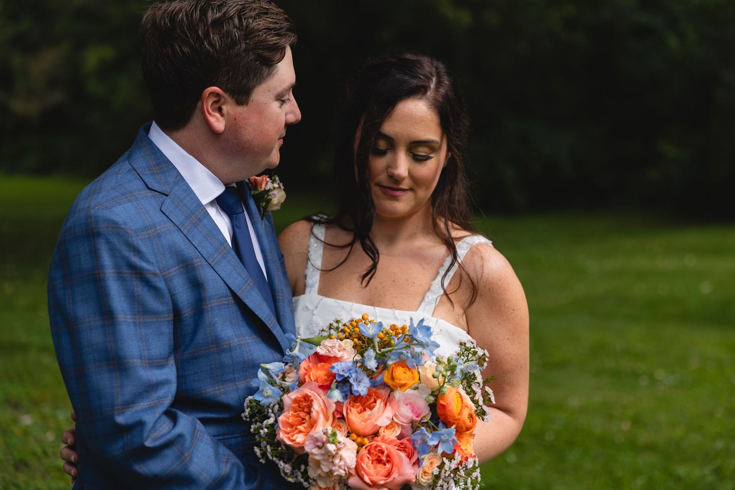 A couple shares a moment on their wedding day with a vibrant coral and blue floral bouquet in a garden setting.