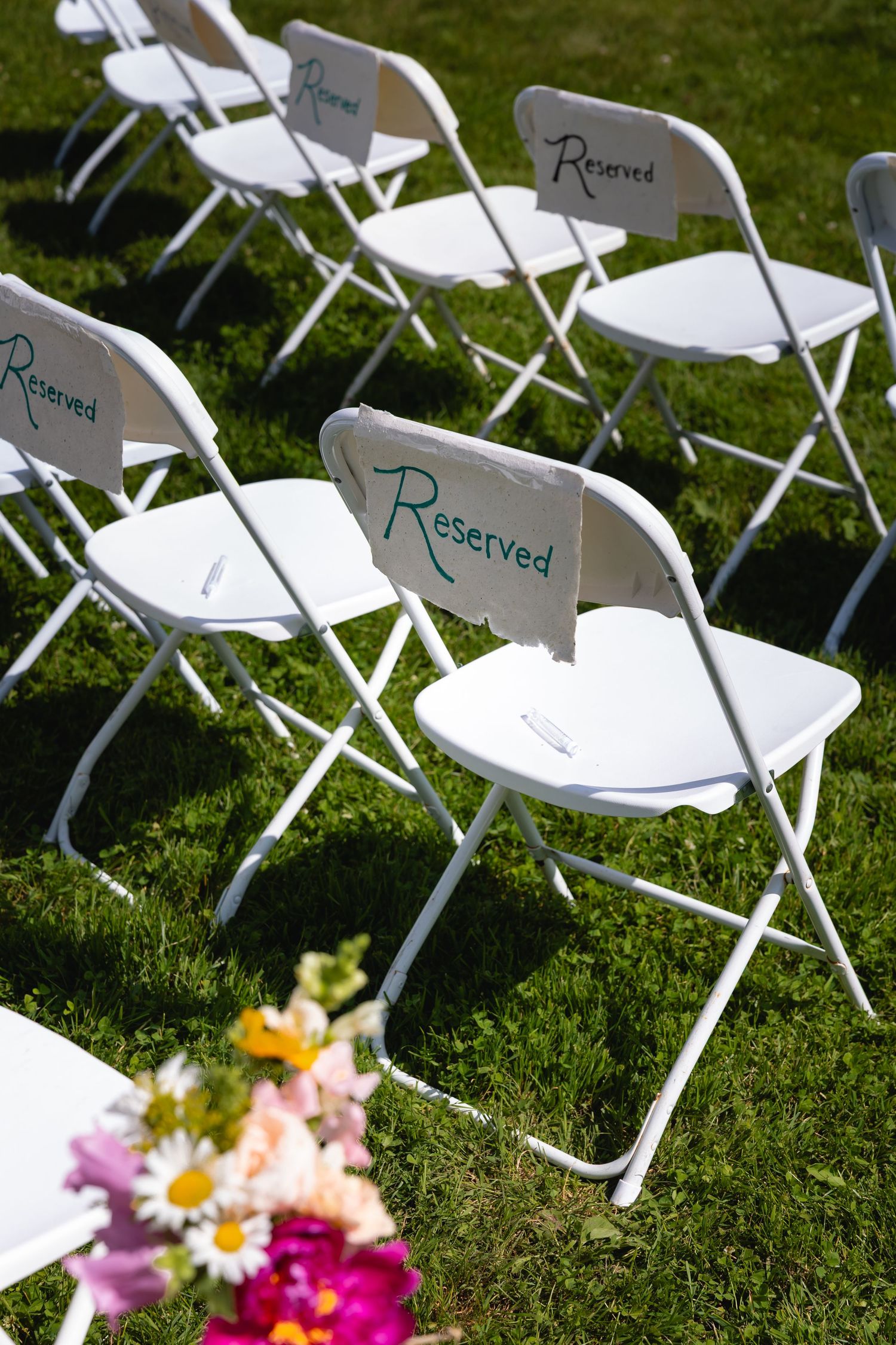 White folding chairs with reserved signs set up on green grass for an outdoor ceremony with colorful floral decorations.