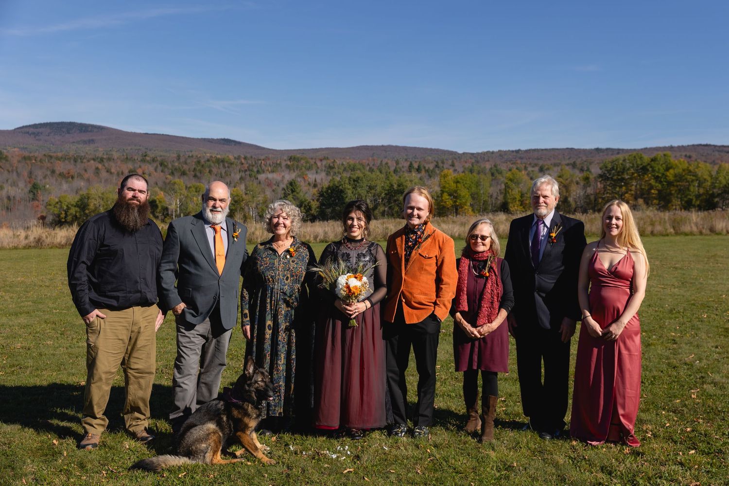A group gathers outdoors for photos in a scenic field with autumn mountains and fall foliage in the background.
