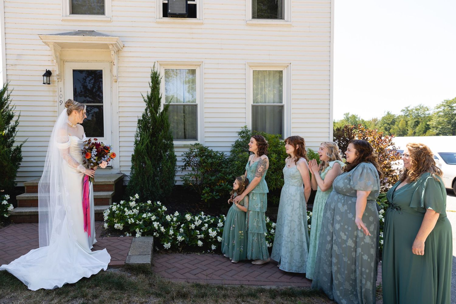 Wedding party members in sage green dresses gather outside a white church building during a summer celebration.