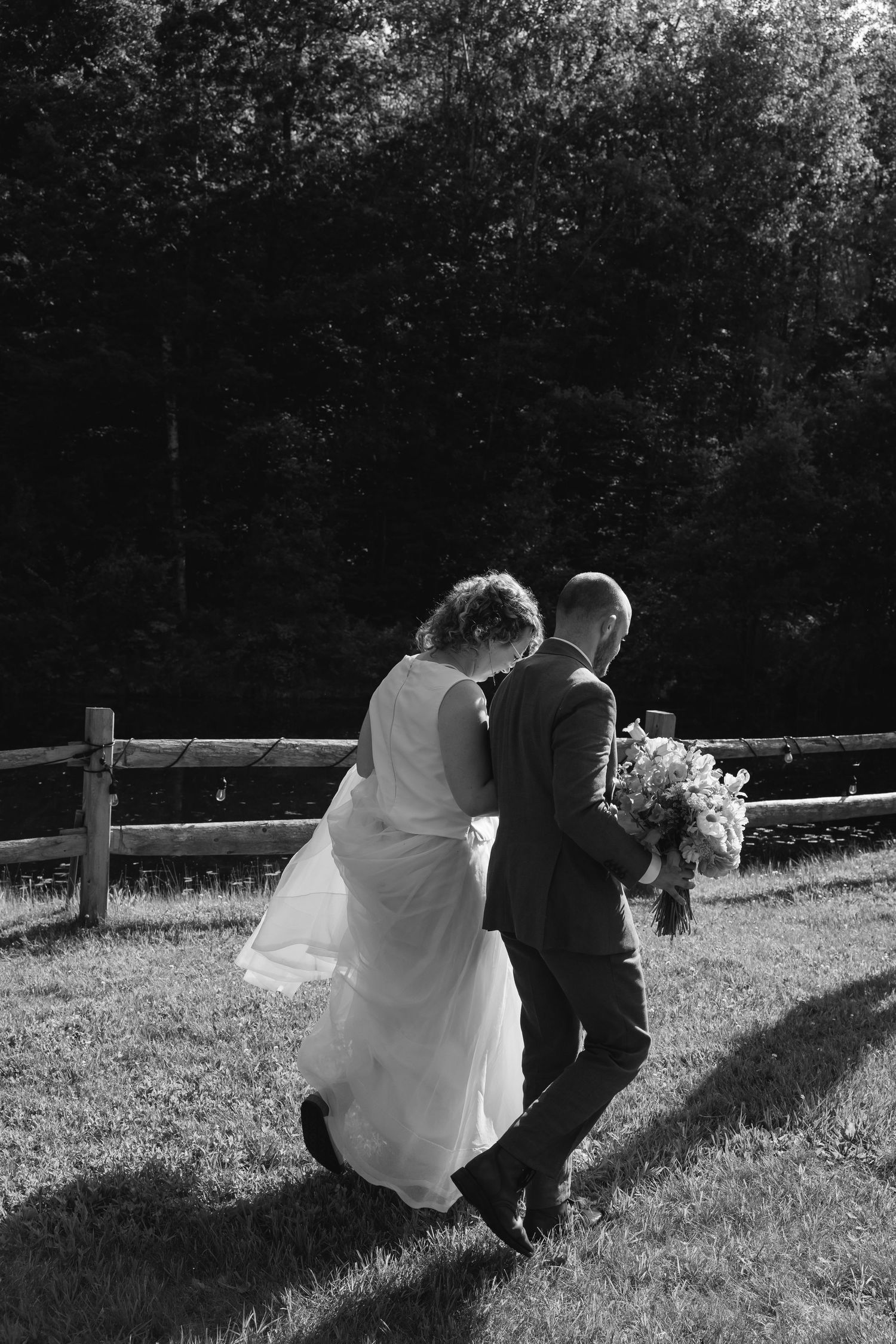 A black and white photo of newlyweds walking together along a rustic fence line holding a wedding bouquet.