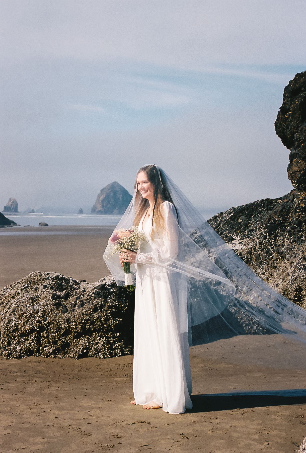 A bride in a flowing white dress and veil stands on a rocky beach with dramatic coastal rock formations in the background.