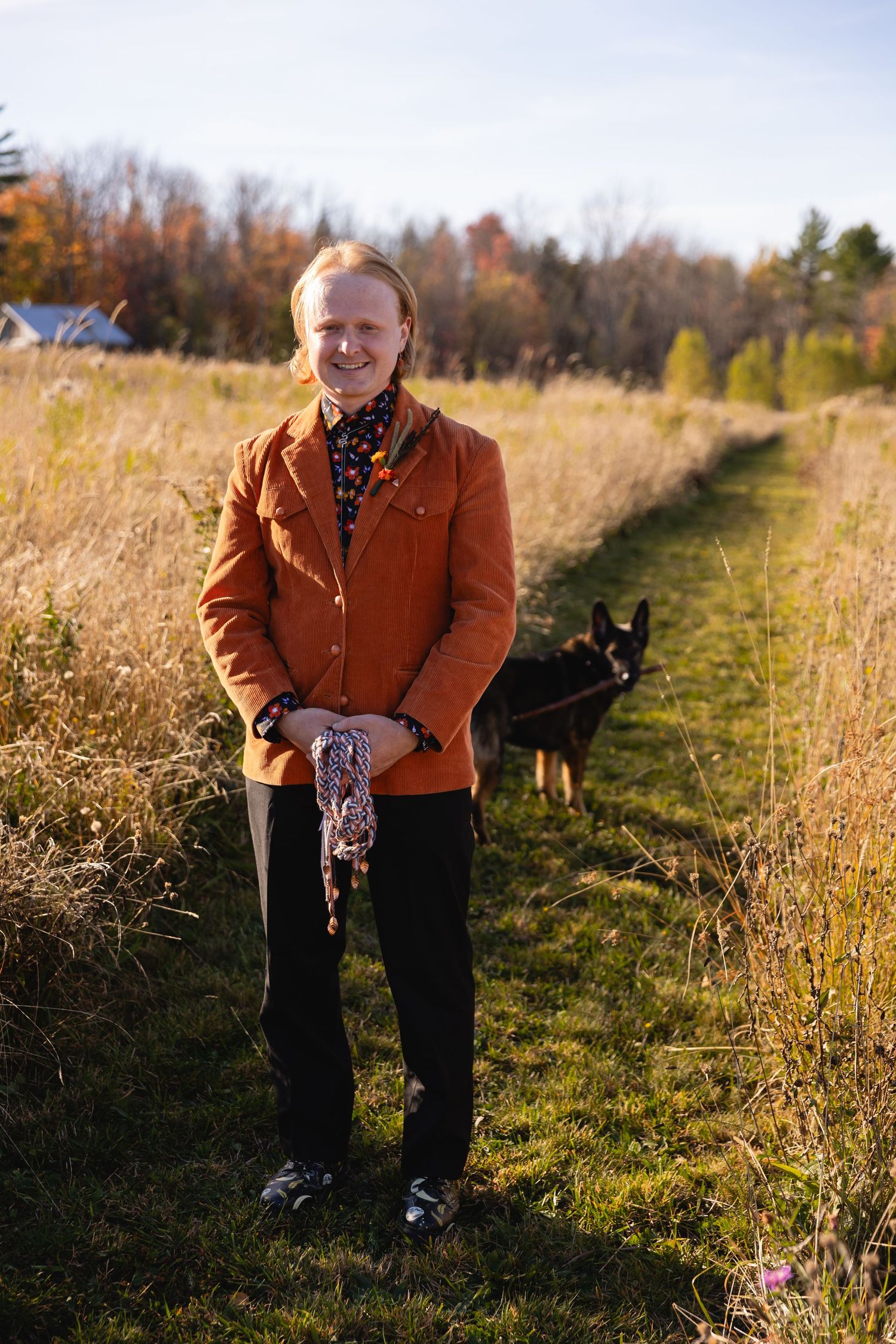 Someone in a brown jacket sits in a grassy field at sunset with a dog visible in the background.