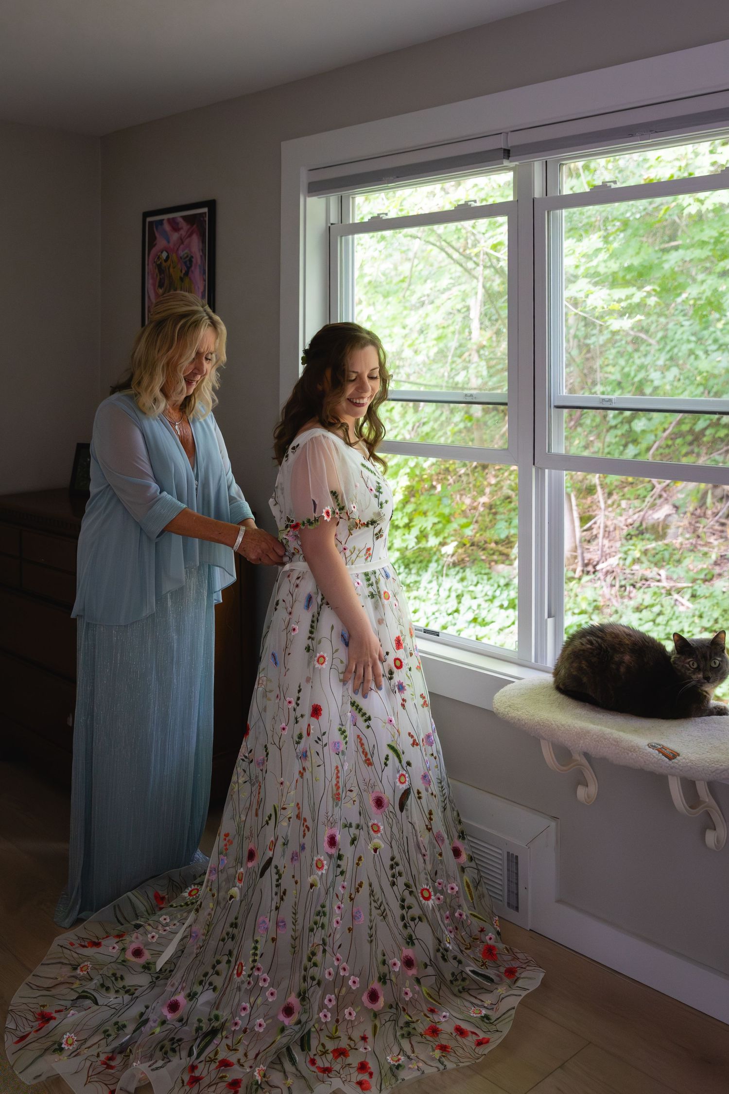 Two people in flowing dresses stand by a window while a black cat lounges on the windowsill in a bright room.