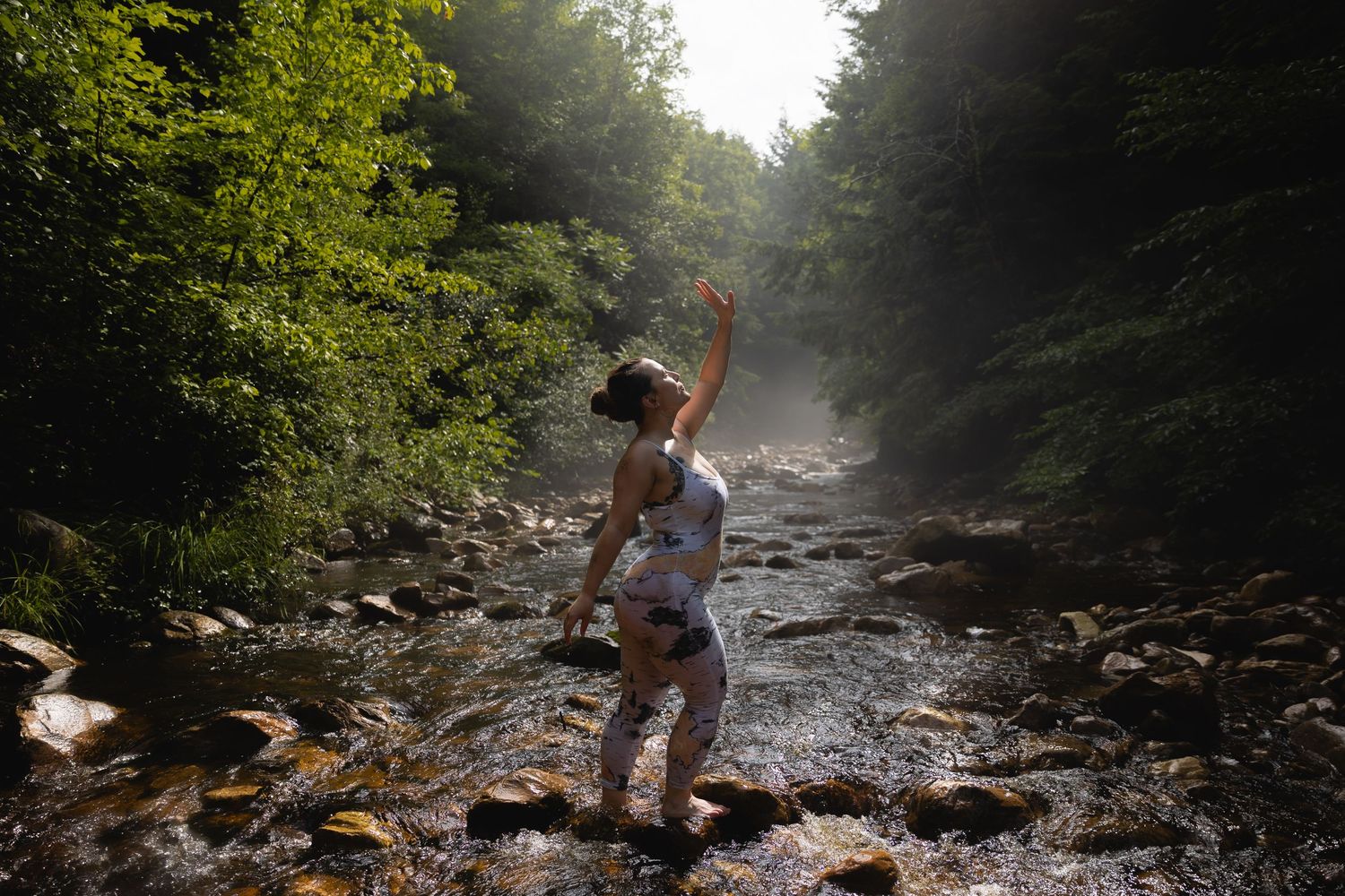 A woman dances joyfully in a sunlit rocky forest stream with arms raised.