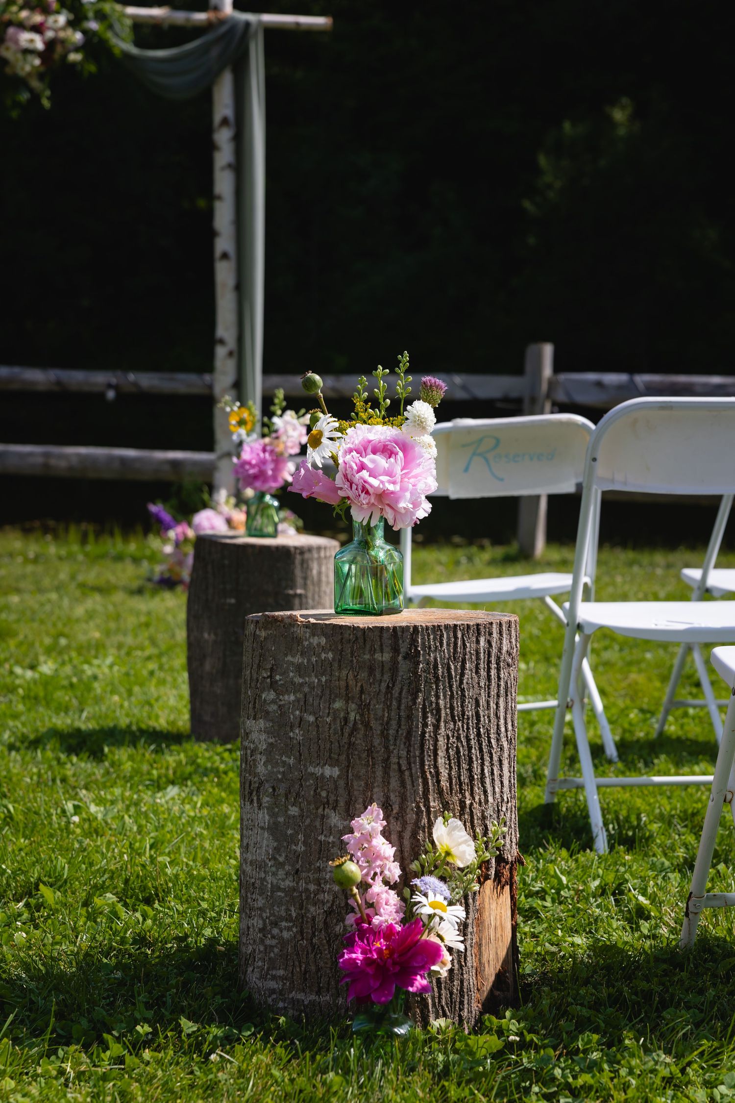 Tree stumps decorated with pink flowers line an outdoor wedding aisle with white folding chairs on green grass.