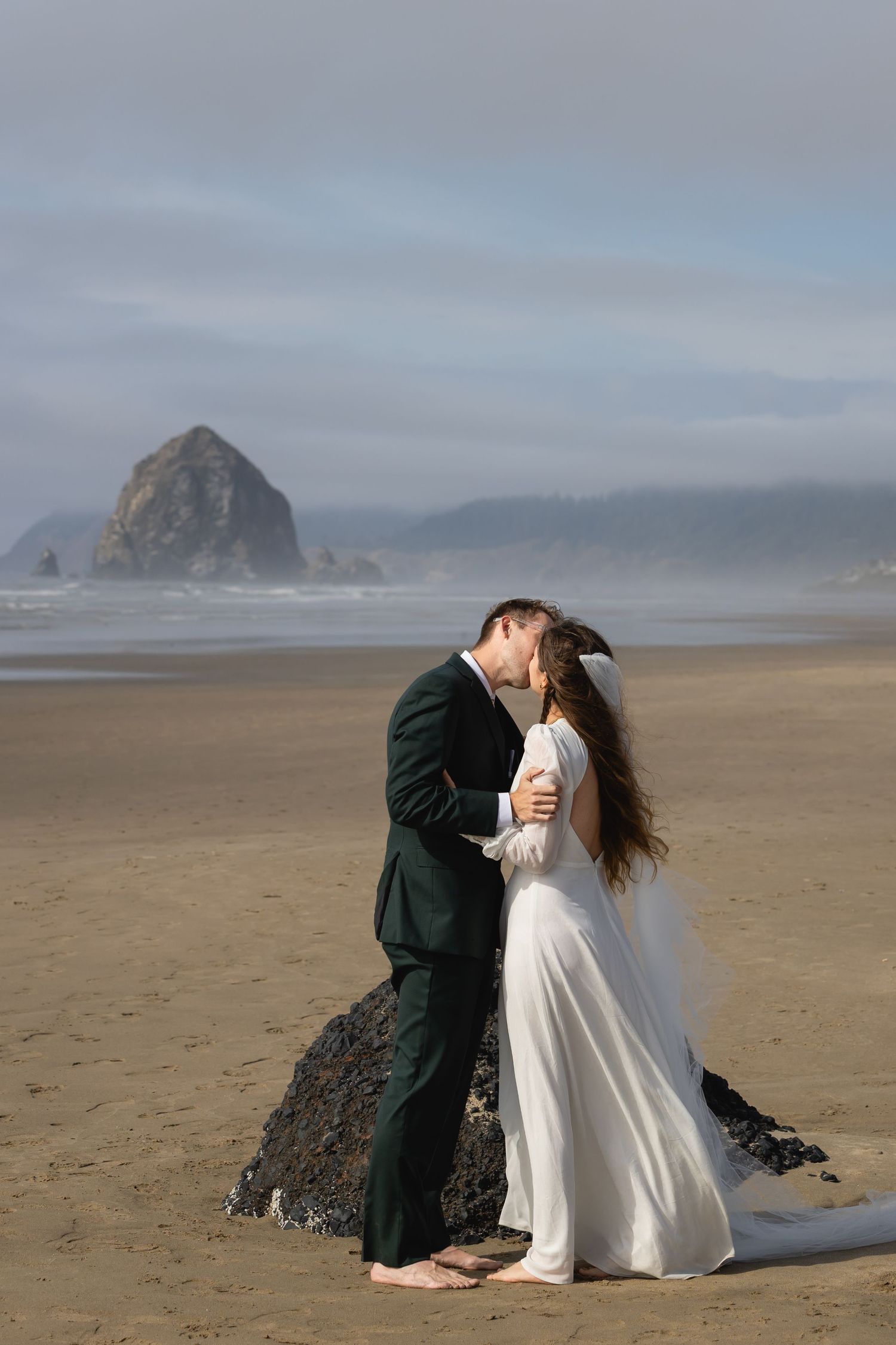 A couple in wedding attire embraces on a misty beach with dramatic coastal rock formations in the background.