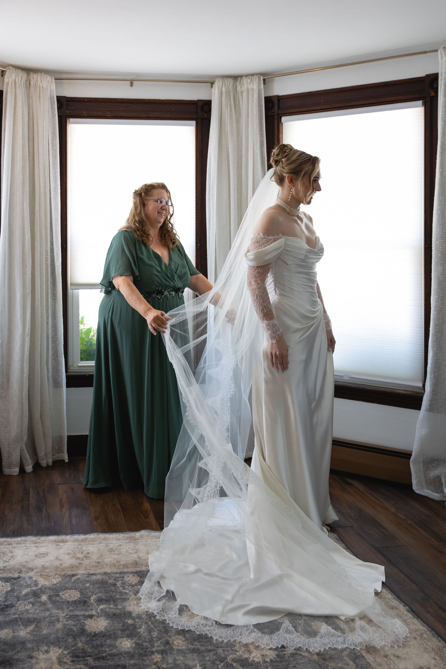 A bride in a flowing white wedding dress gets help with her long veil on a hardwood floor with natural lighting.