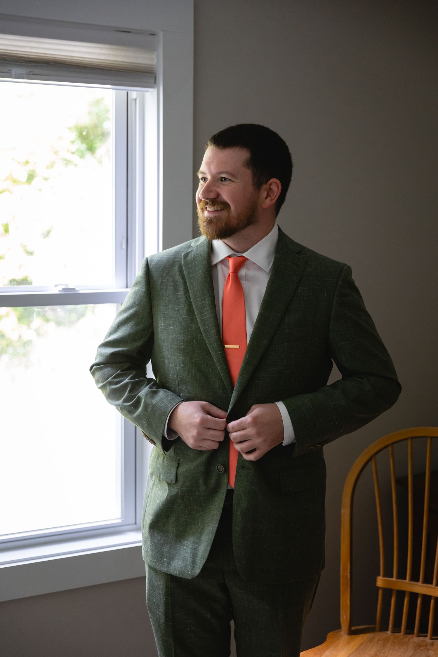 A person in a gray suit and coral tie adjusts their jacket while standing near a window in an indoor setting.