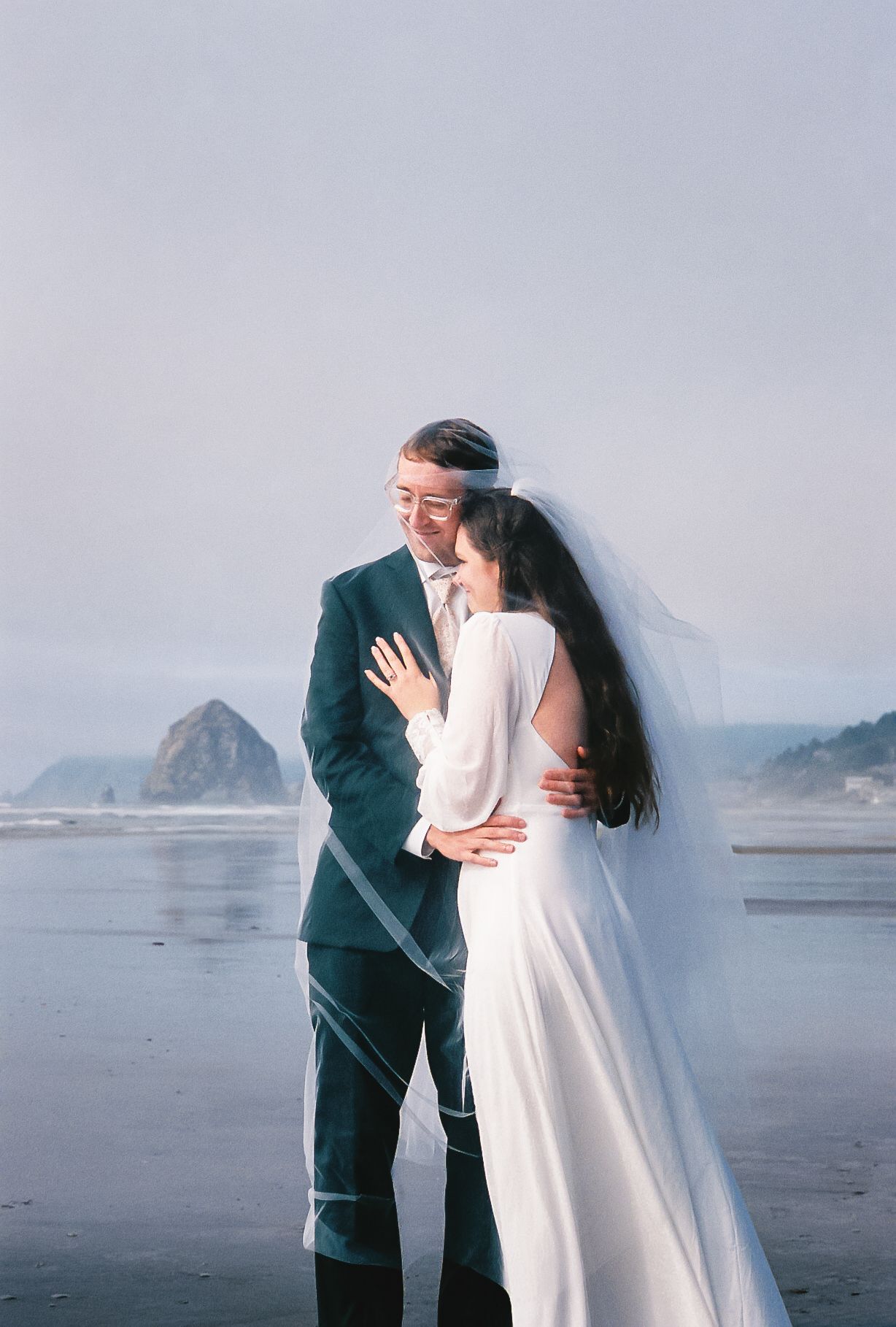A couple in wedding attire shares romantic moments on a misty beach with coastal rock formations in the background.