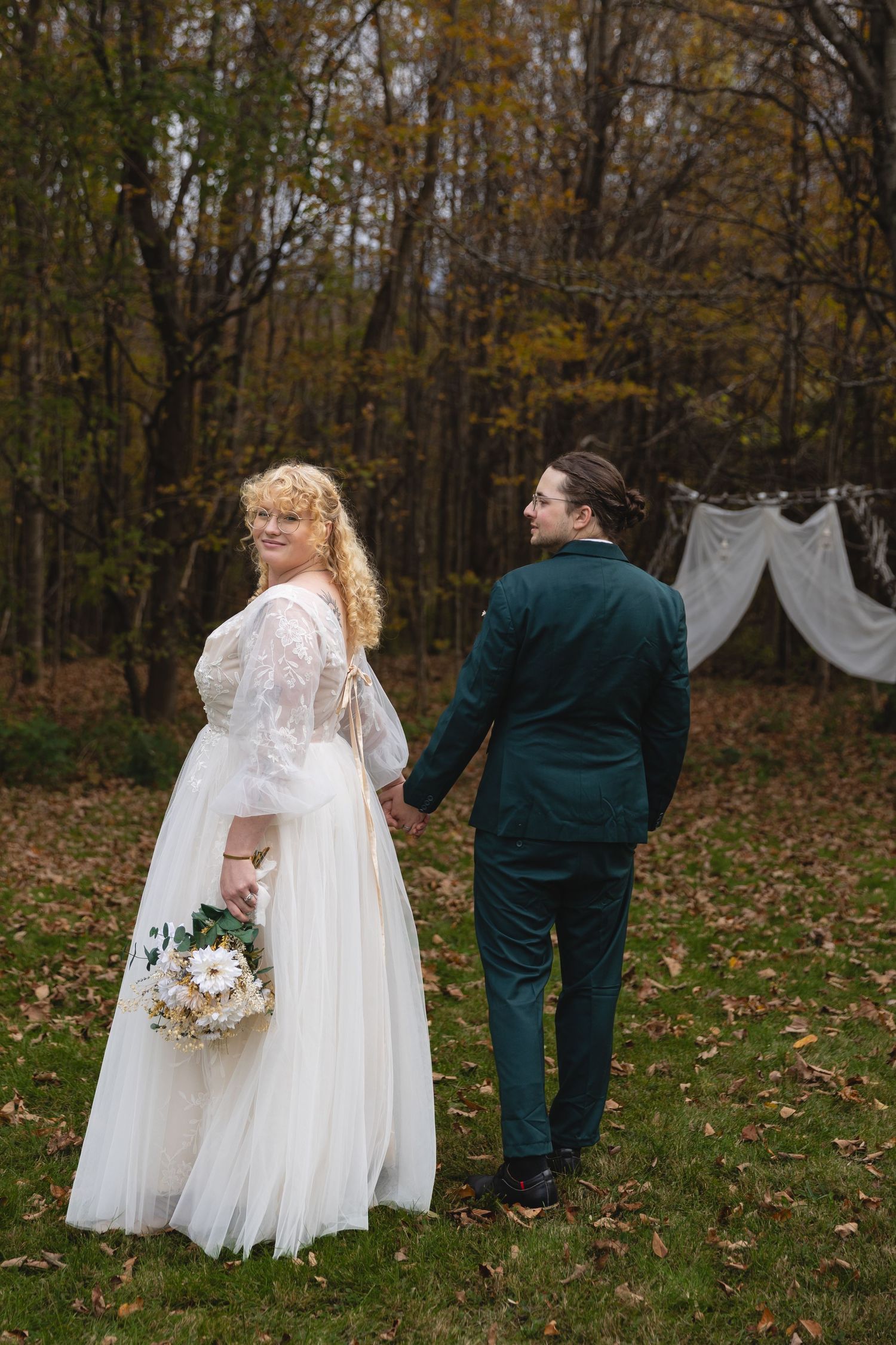 Couple in wedding attire standing in front of white fabric backdrop in autumn woods.