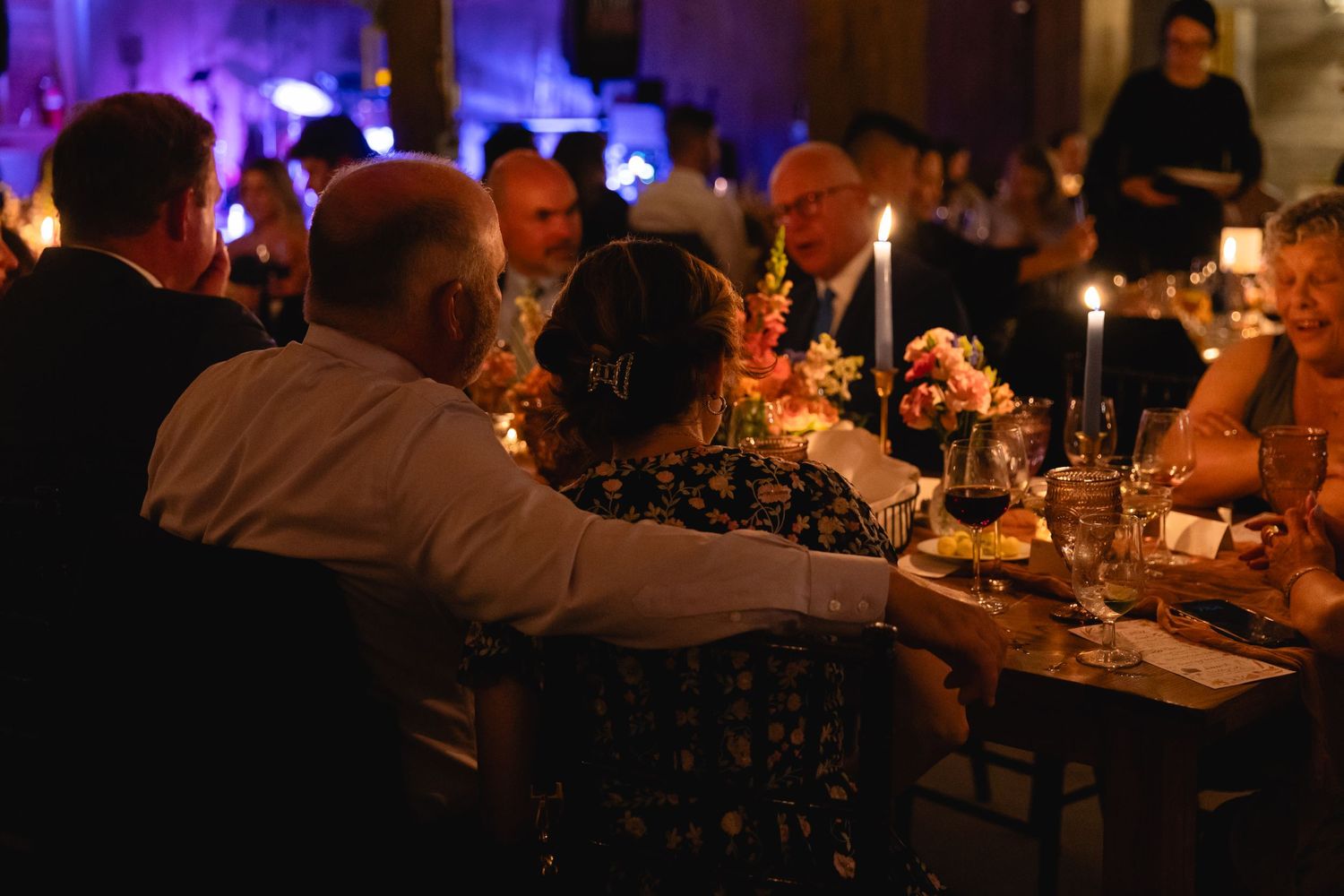 Wedding guests seated at a candlelit table during an elegant evening reception with purple ambient lighting.